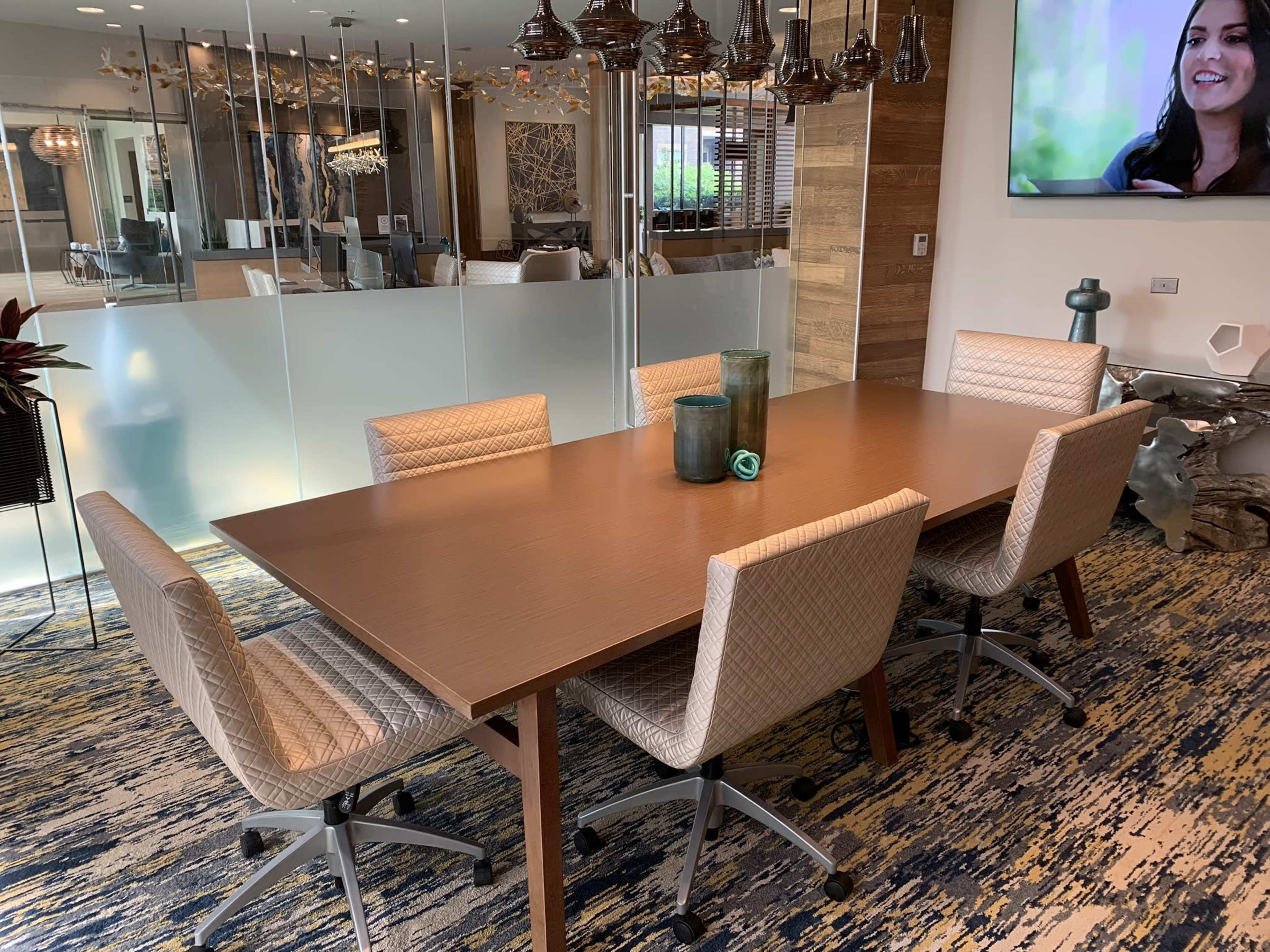 A conference room features a wooden table surrounded by five upholstered chairs, with decorative vases in the center and a television mounted on the wall.