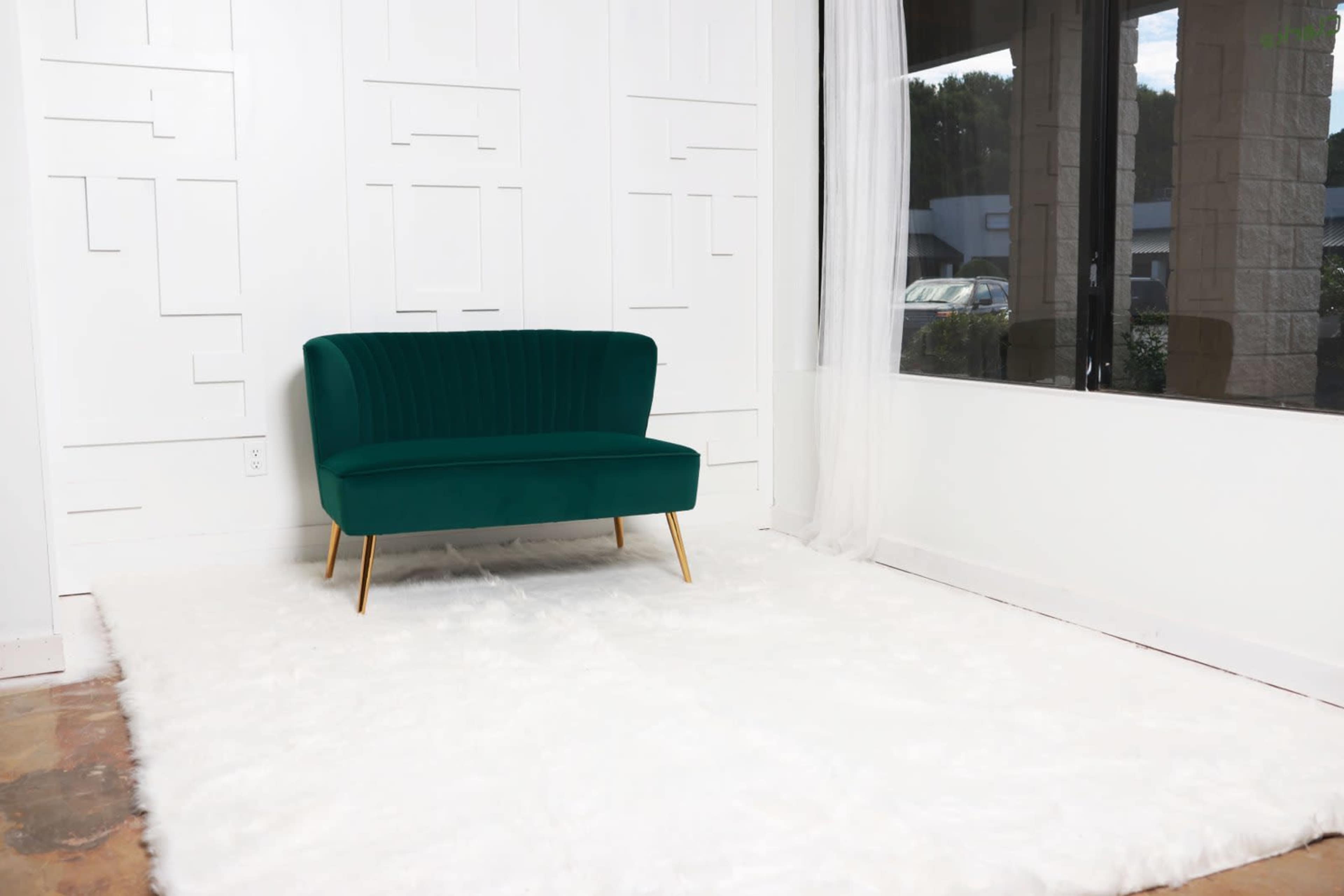The image shows a green velvet loveseat on a white, fluffy rug in a room with a large window and a textured white wall.