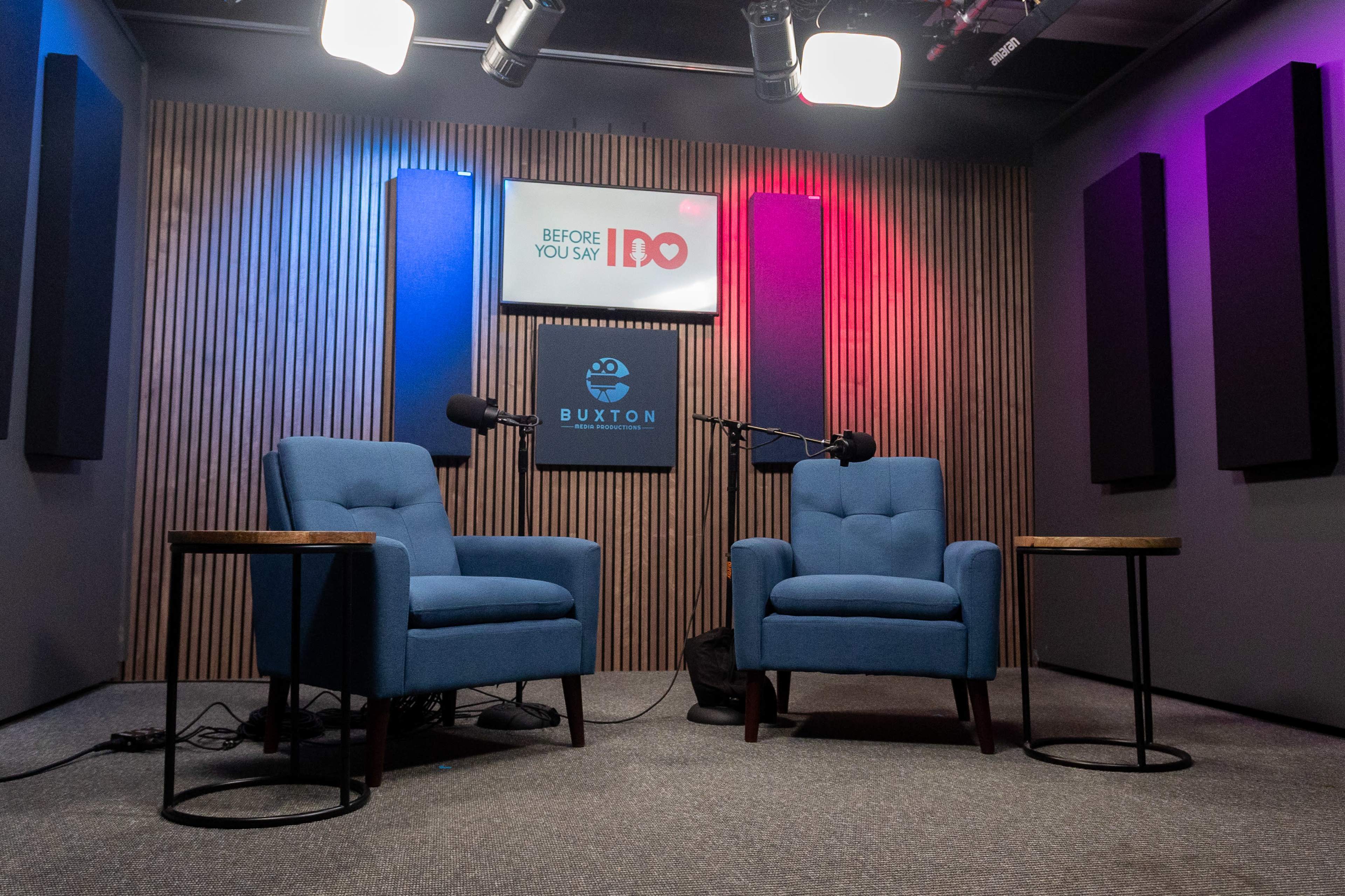 The image shows a podcast recording studio with two blue chairs, a microphone on each side, and colorful lighting in the background.