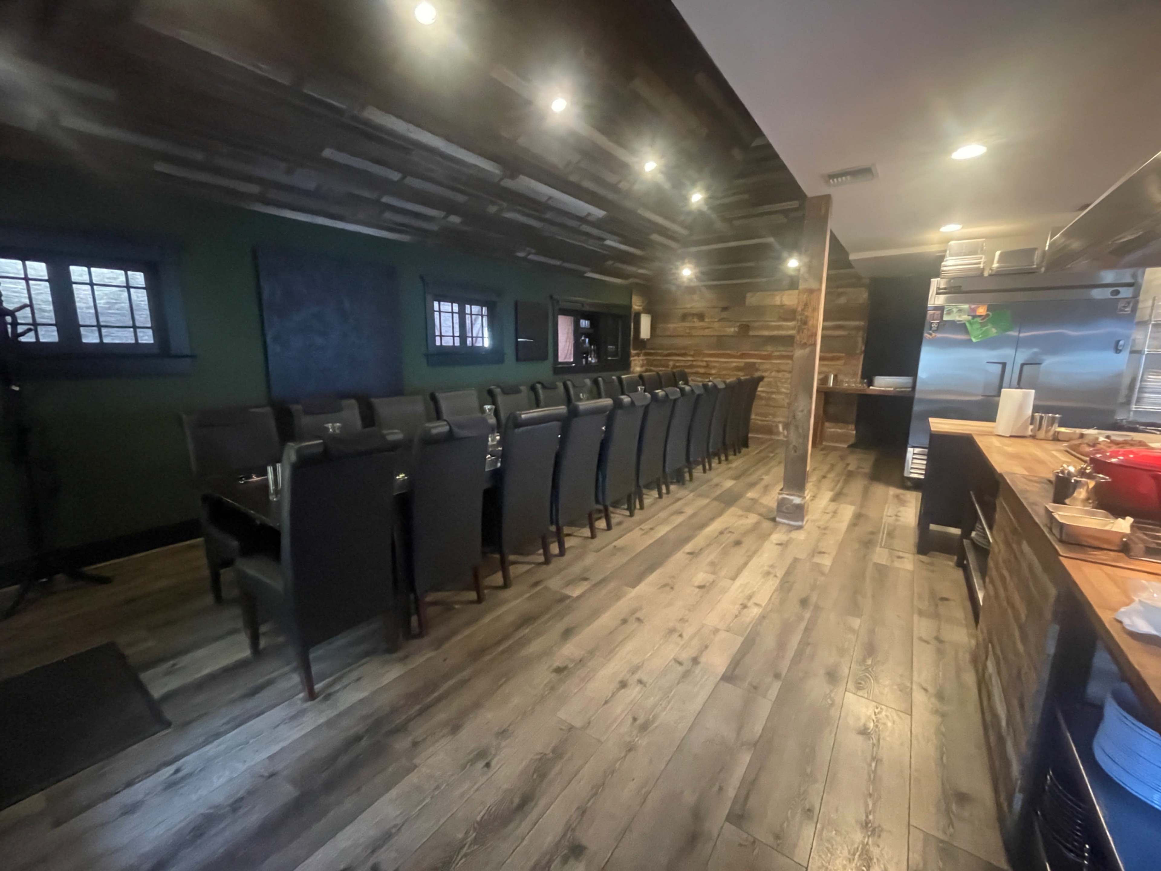 The image shows a modern restaurant interior with a long row of black chairs and a wooden bar area, illuminated by overhead lighting.