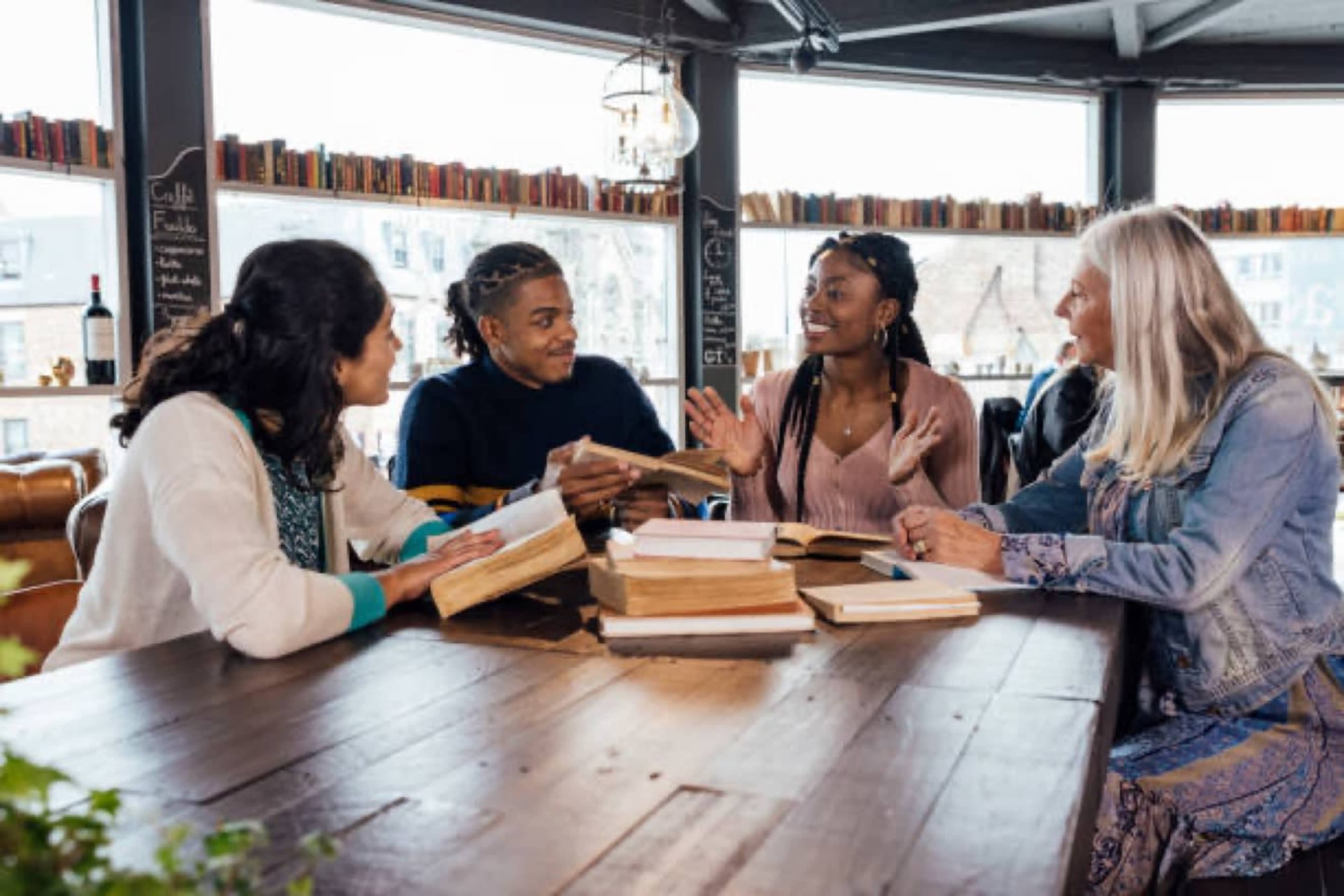 A group of four people is seated around a wooden table, engaged in conversation while surrounded by books in a cozy reading space.