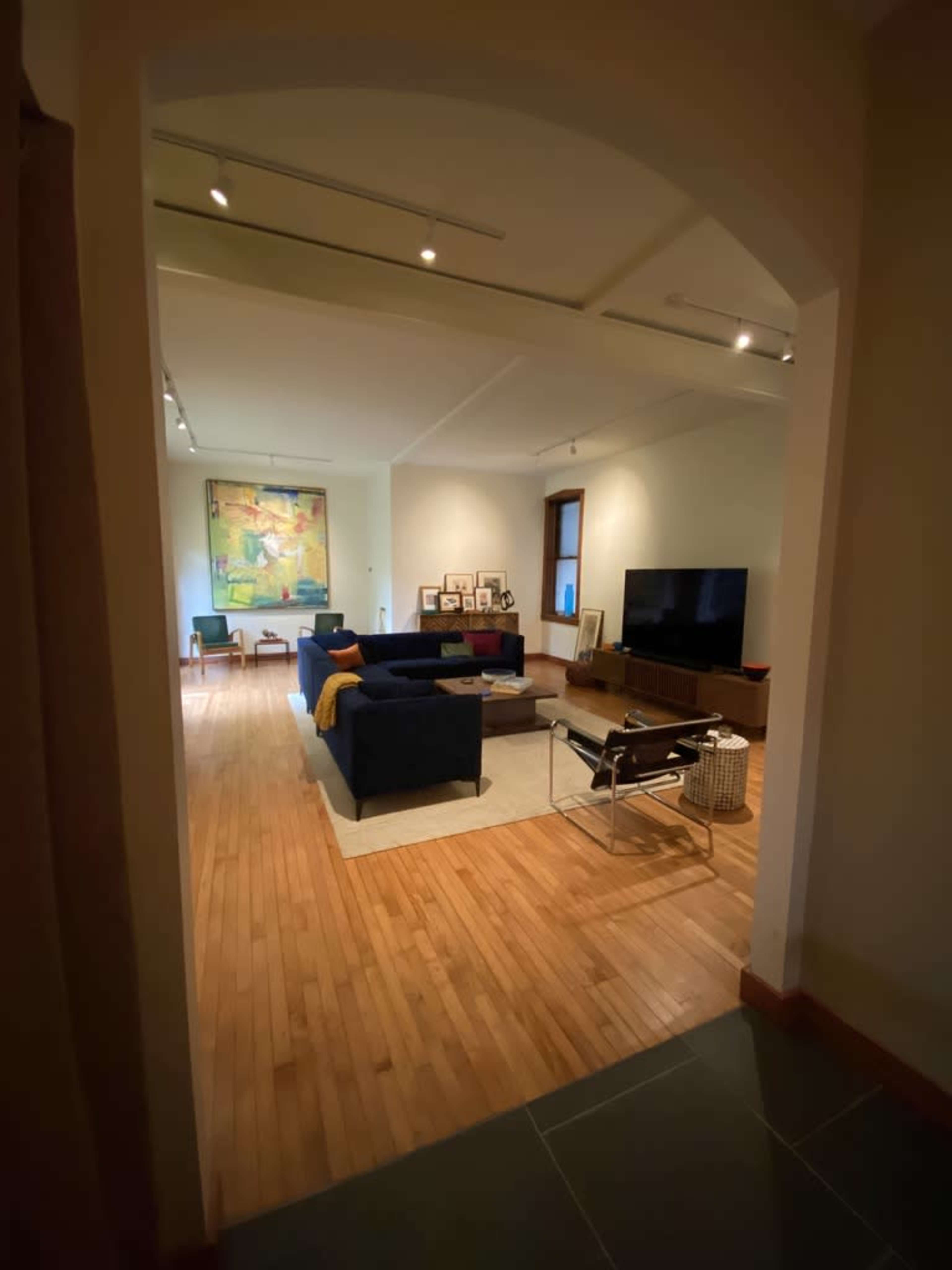 The image shows a spacious living area with wooden flooring, a large television, comfortable seating, and an abstract painting on the wall.