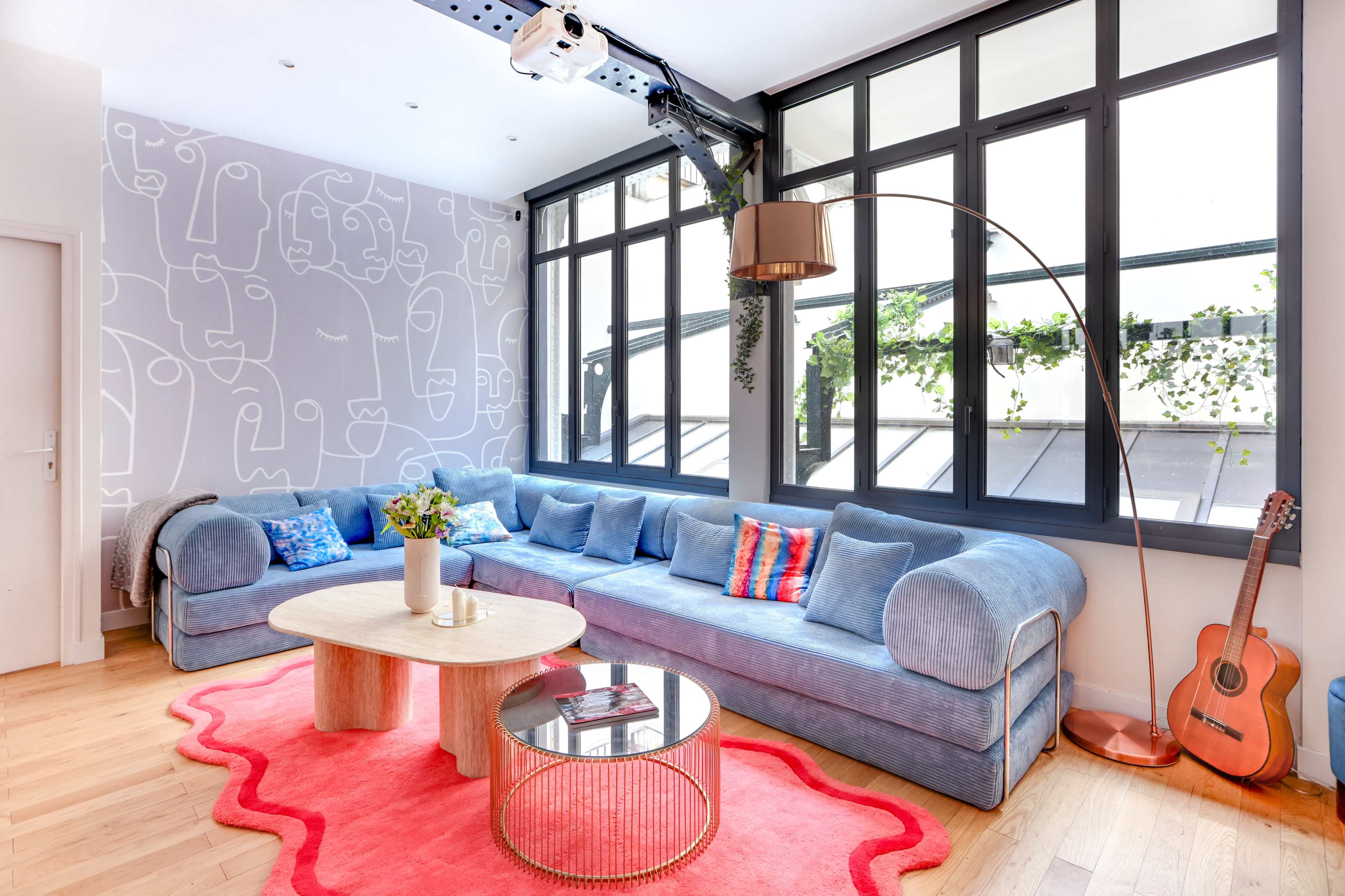 A modern living room features a blue couch, a circular coffee table, and large windows letting in natural light, with a guitar and vibrant decor elements.
