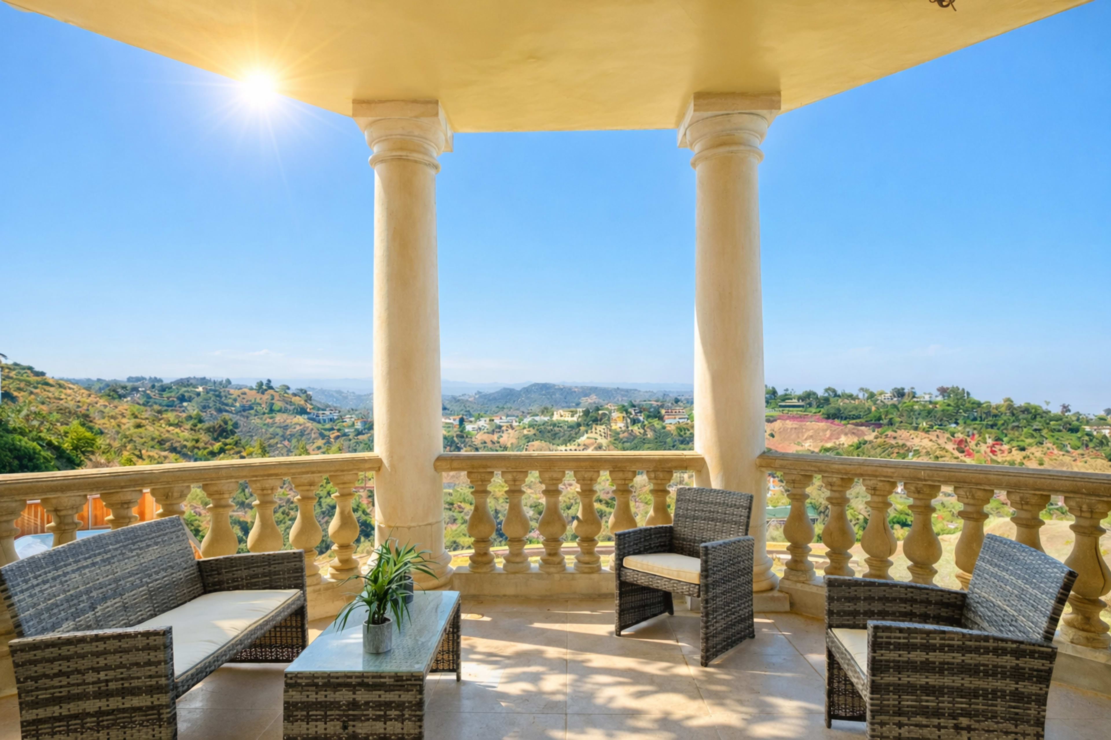 A spacious balcony features woven furniture and offers a panoramic view of rolling hills under a clear blue sky.