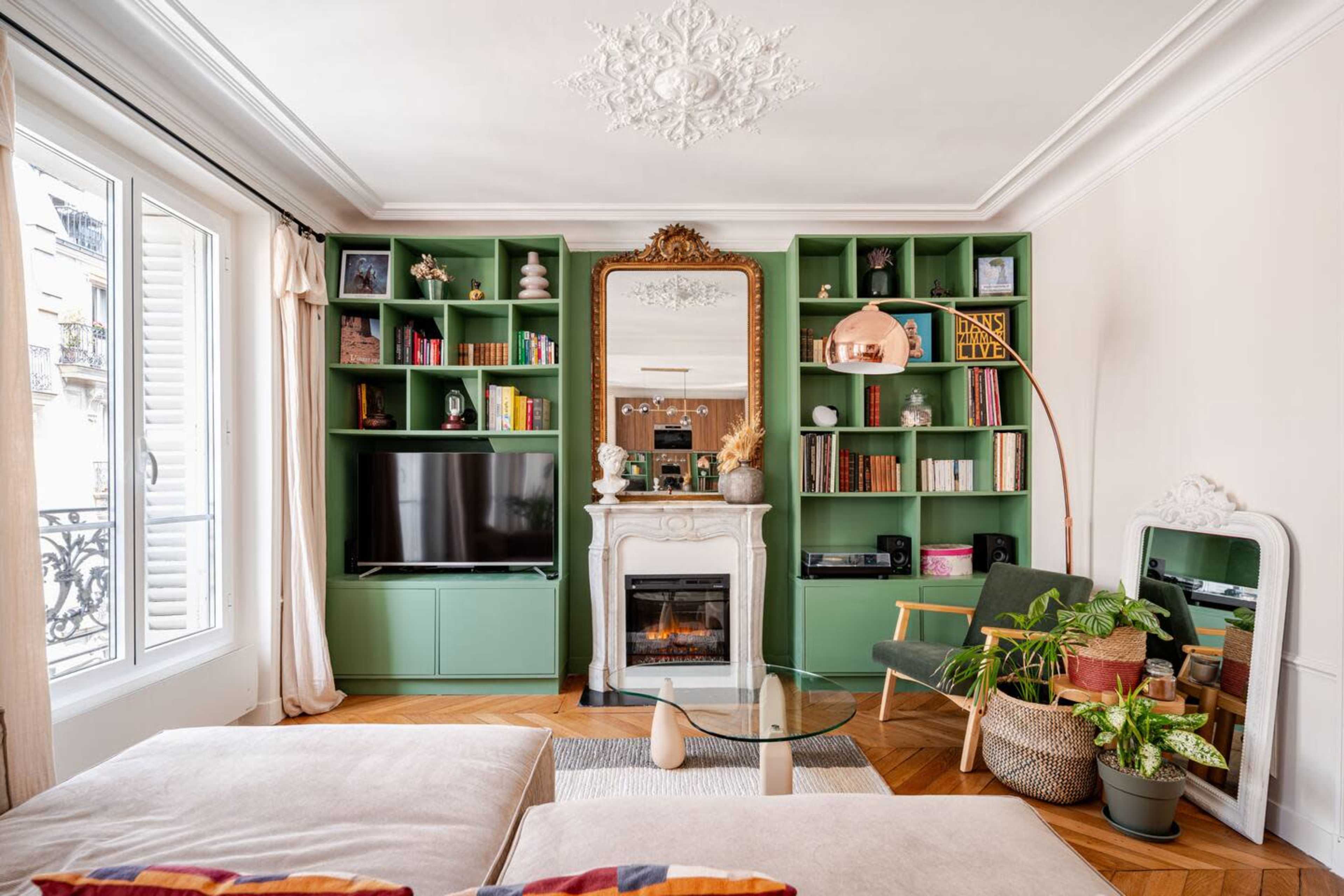 The living room features a green bookshelf with decorative items, a TV, a white fireplace, and comfortable seating, illuminated by natural light from the window.