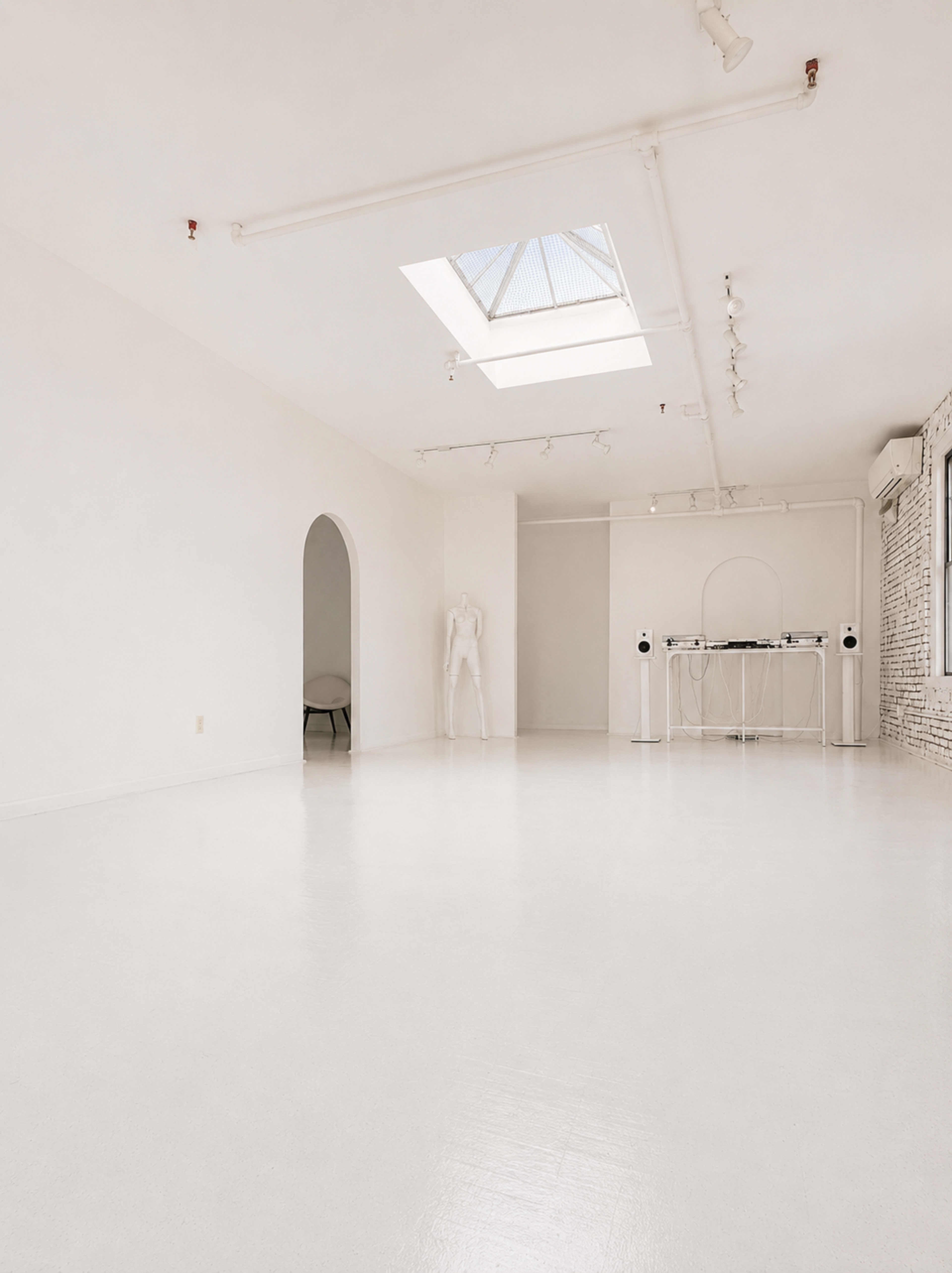 The image shows a spacious, minimalistic interior featuring white walls, a skylight, and a small stage with audio equipment.