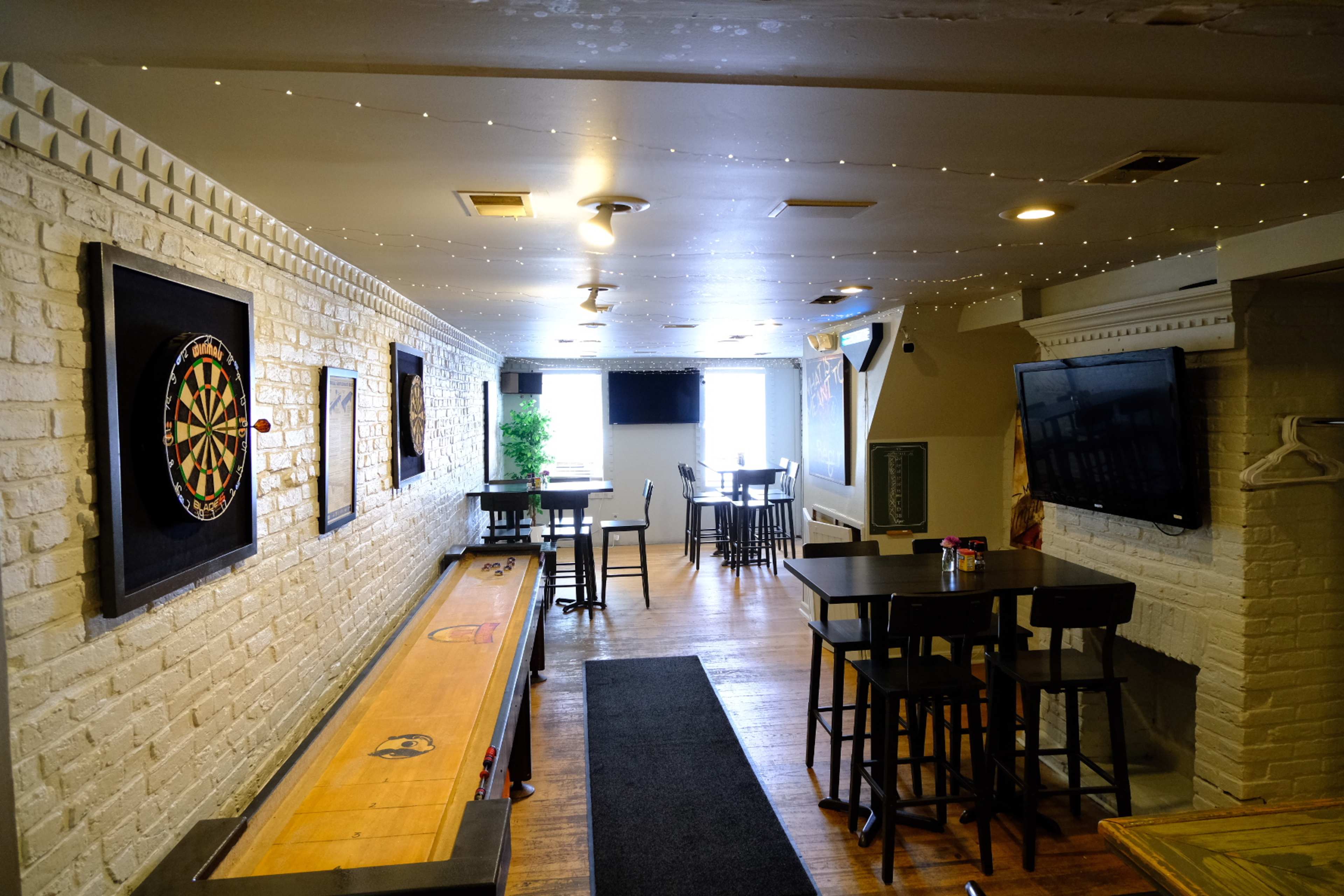 A narrow, well-lit bar area featuring a shuffleboard, dartboard, and tables with high stools against white brick walls.