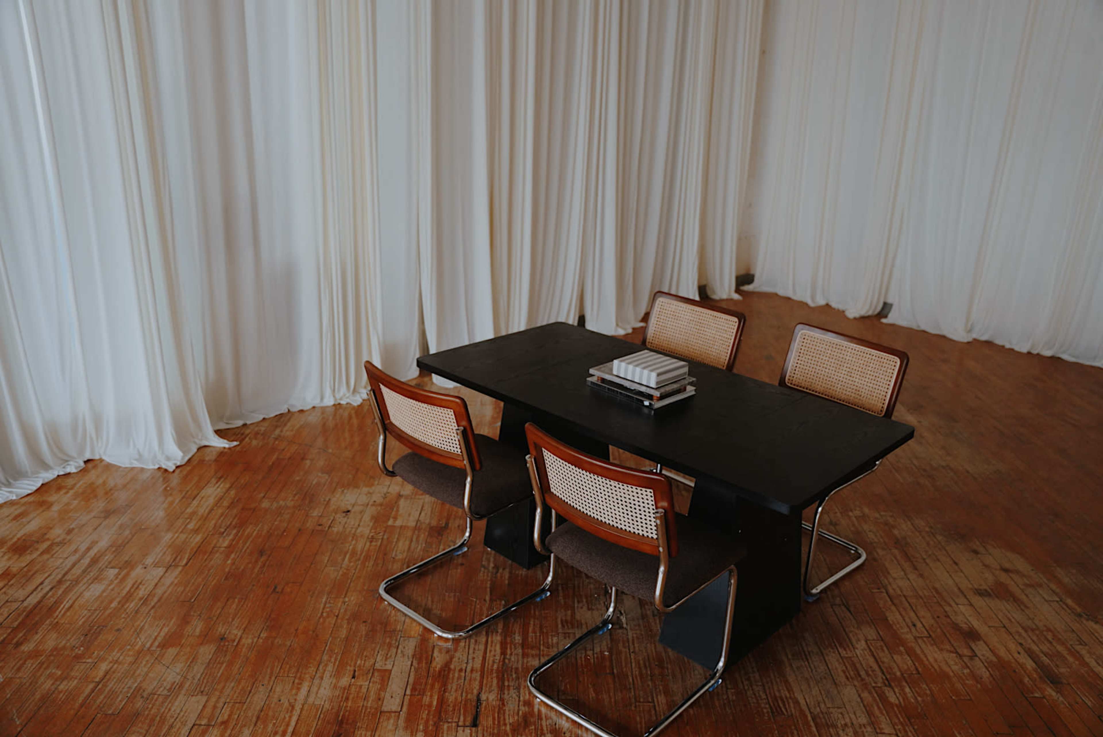 A wooden table surrounded by four chairs is set in a room with white draped fabric and wooden flooring.