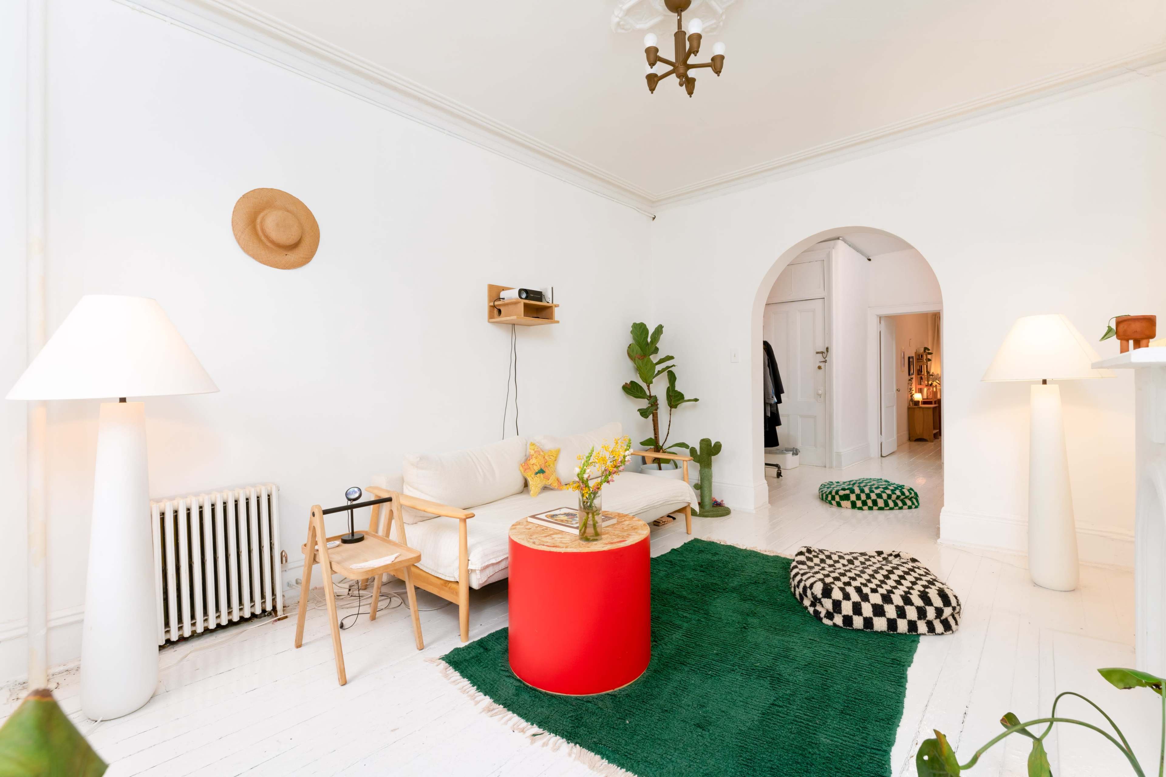 A modern living room features a white sofa, a red coffee table, two floor lamps, a potted plant, and patterned cushions on a green rug.