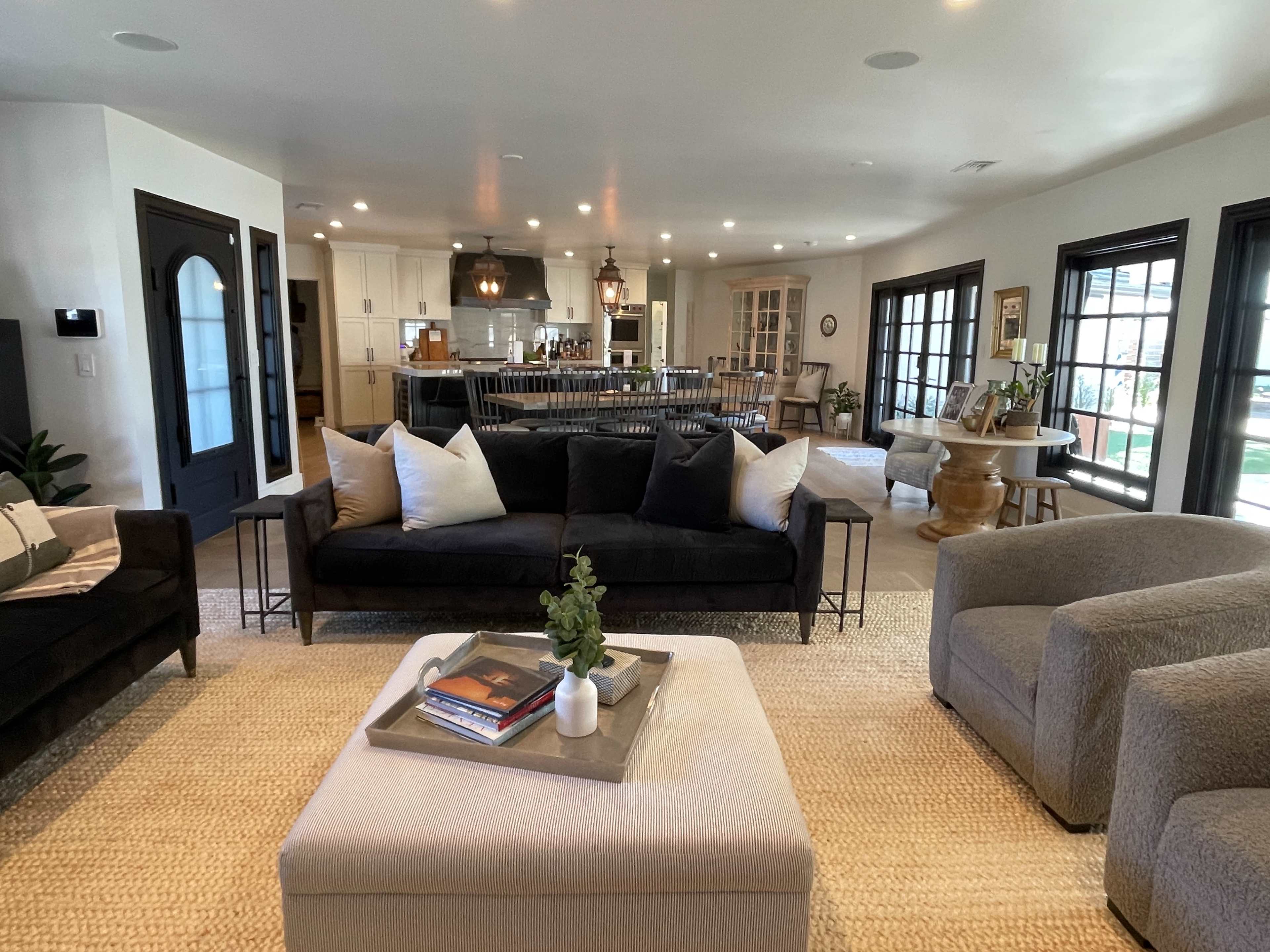 A spacious living area with a black sofa, light-colored armchairs, a coffee table, and a view of a kitchen with wooden cabinetry in the background.