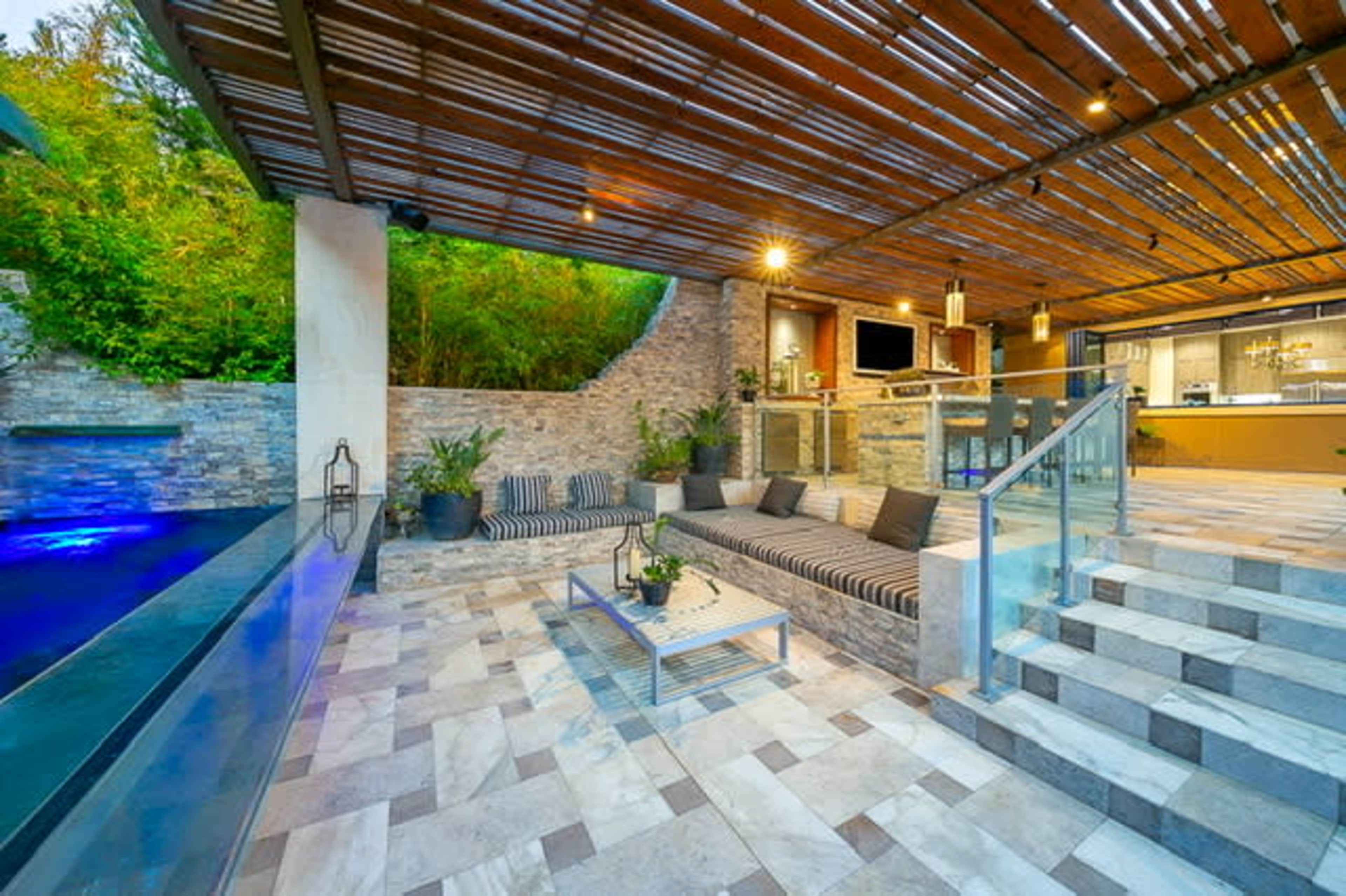 A modern outdoor living space with a lounge area, a stone wall, a swimming pool, and stairs leading to an indoor kitchen.