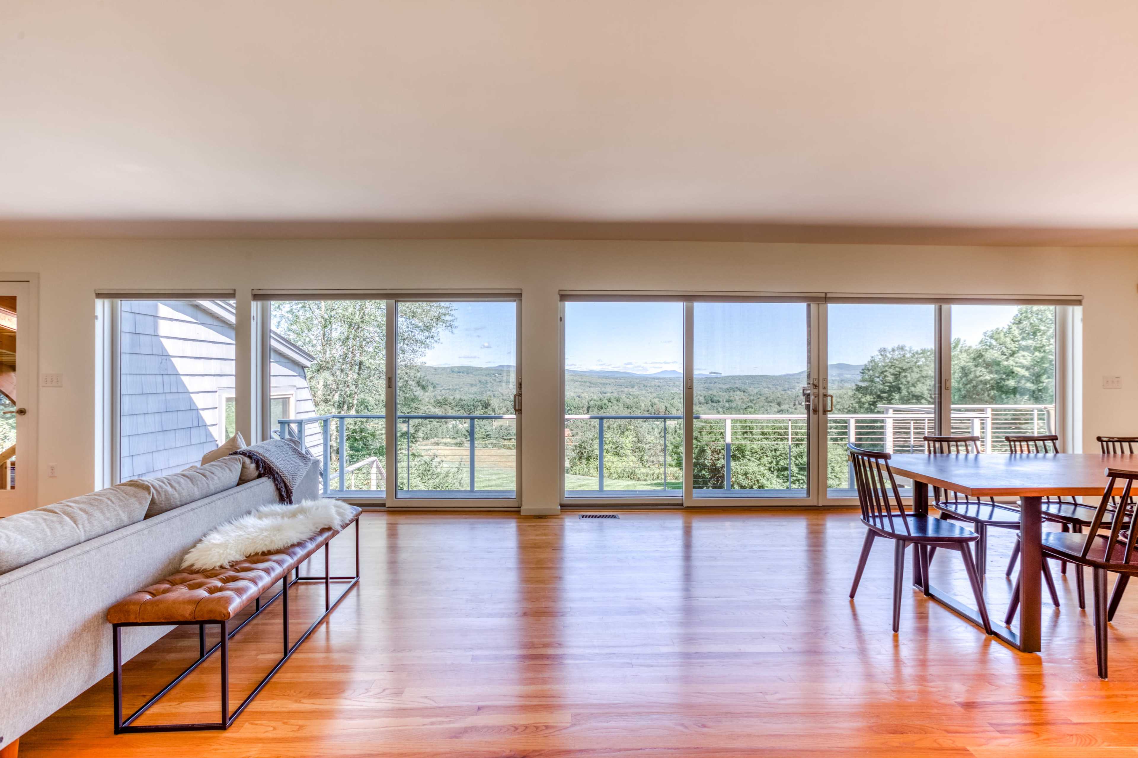 A spacious living room features large windows overlooking a picturesque outdoor landscape, with a sofa and dining table inside.