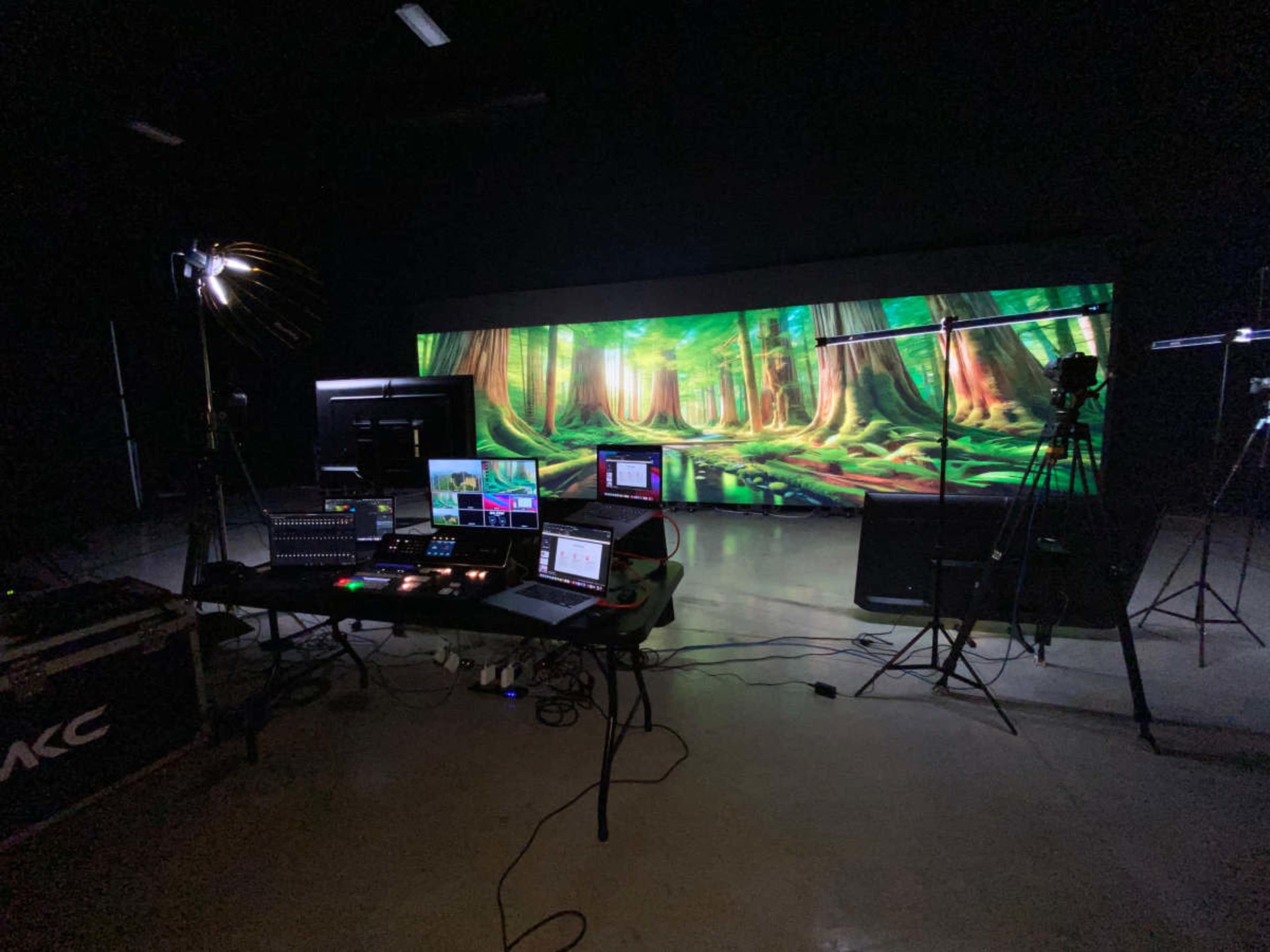 A studio setup with multiple computer screens, cameras, and lighting equipment in front of a large backdrop depicting a forest scene.