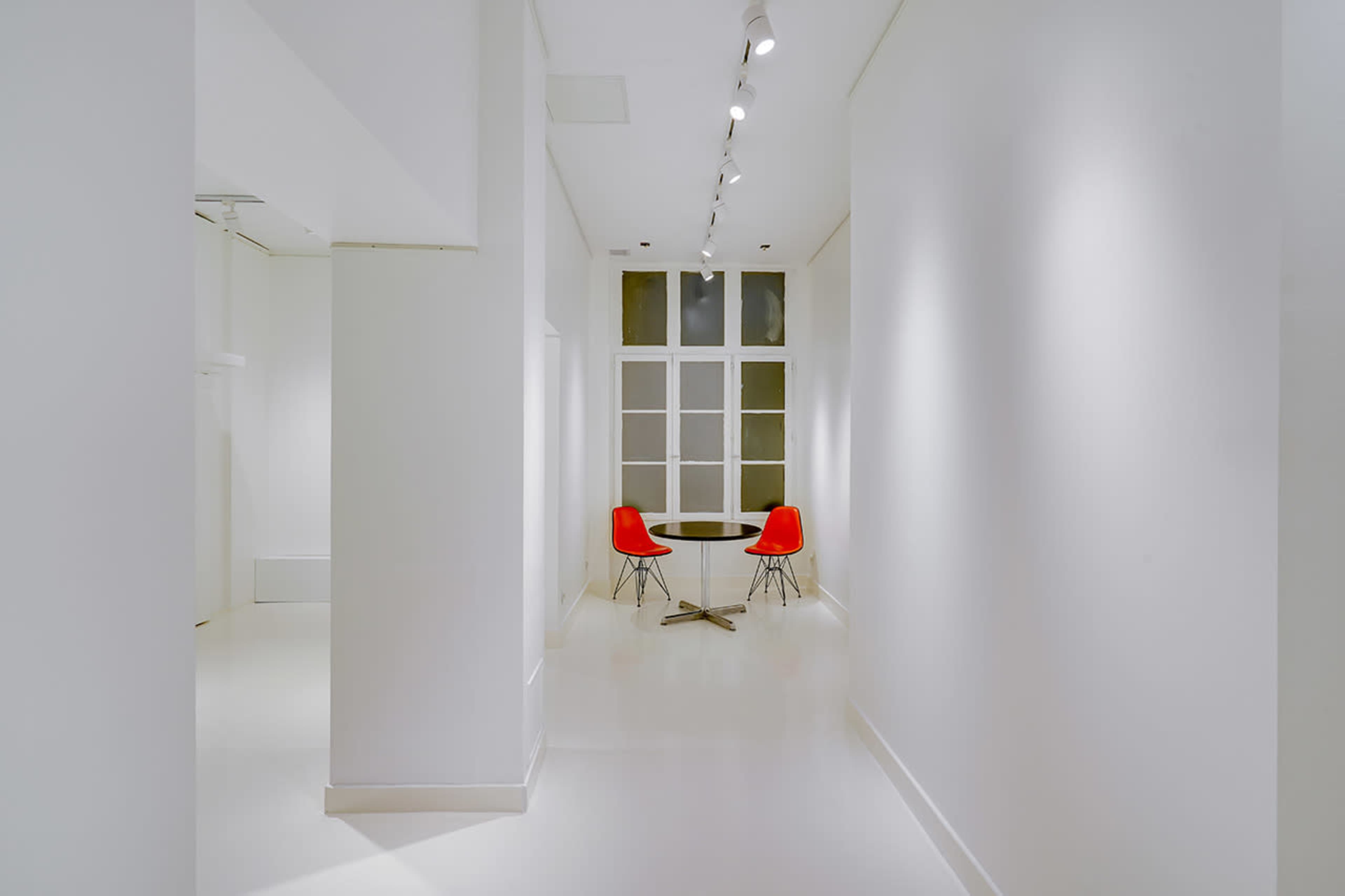 A narrow, brightly lit corridor leads to a small table with two red chairs set against a white backdrop.