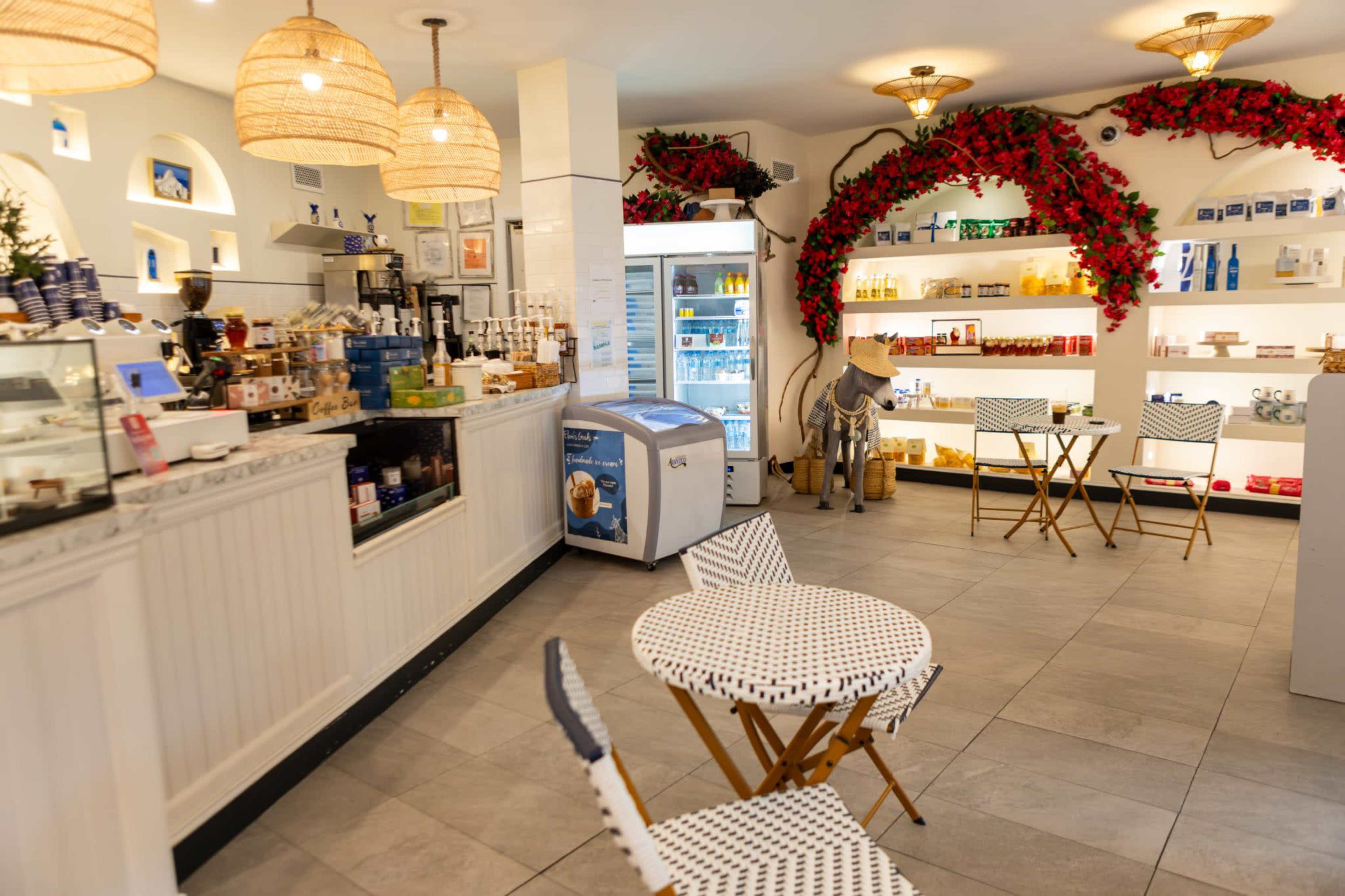 The image shows a bright café interior with a counter displaying various products, a small seating area with two tables, and decorative floral arrangements along the walls.