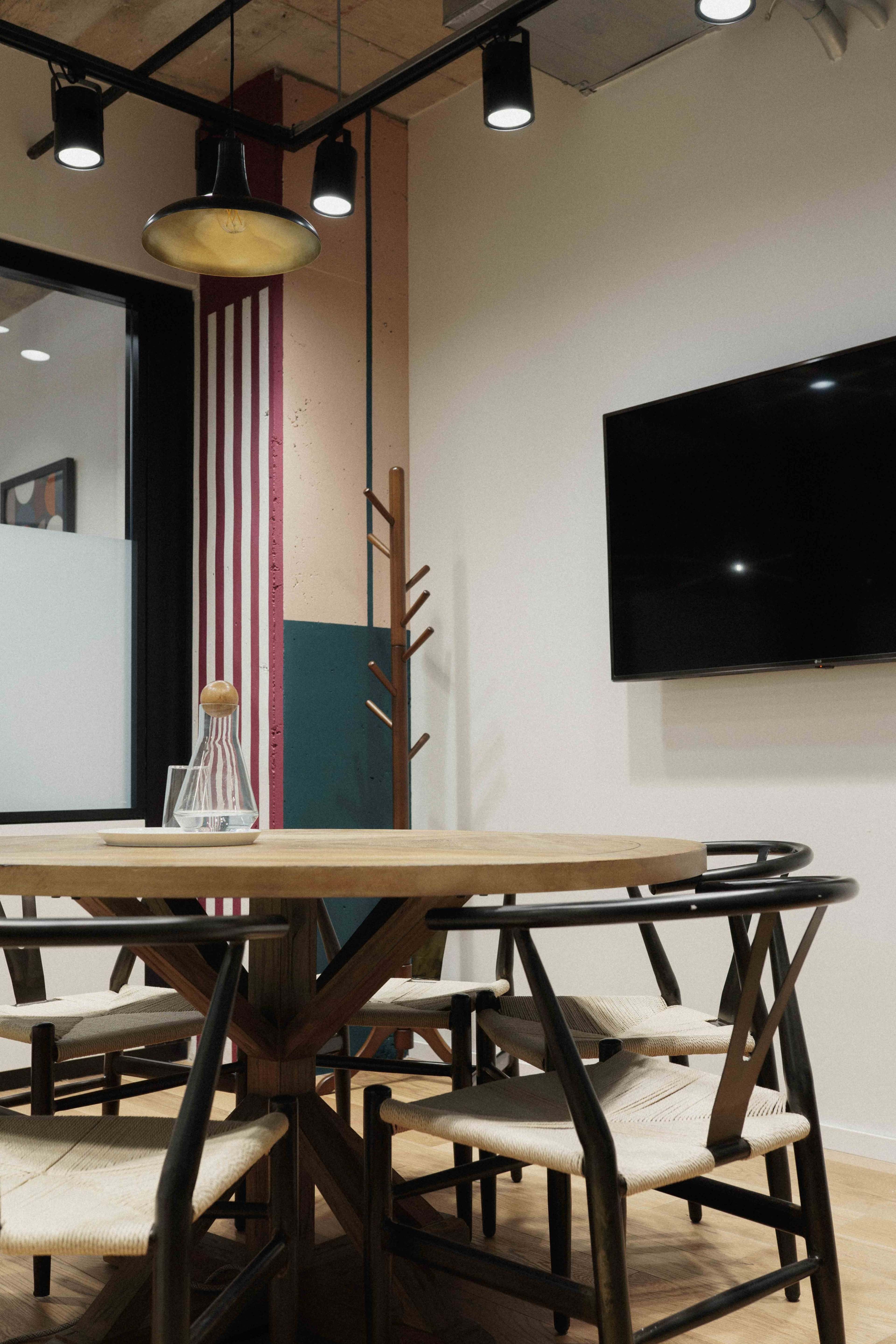 A round wooden table with woven chairs is positioned in a modern office space featuring a wall-mounted TV and a coat rack.