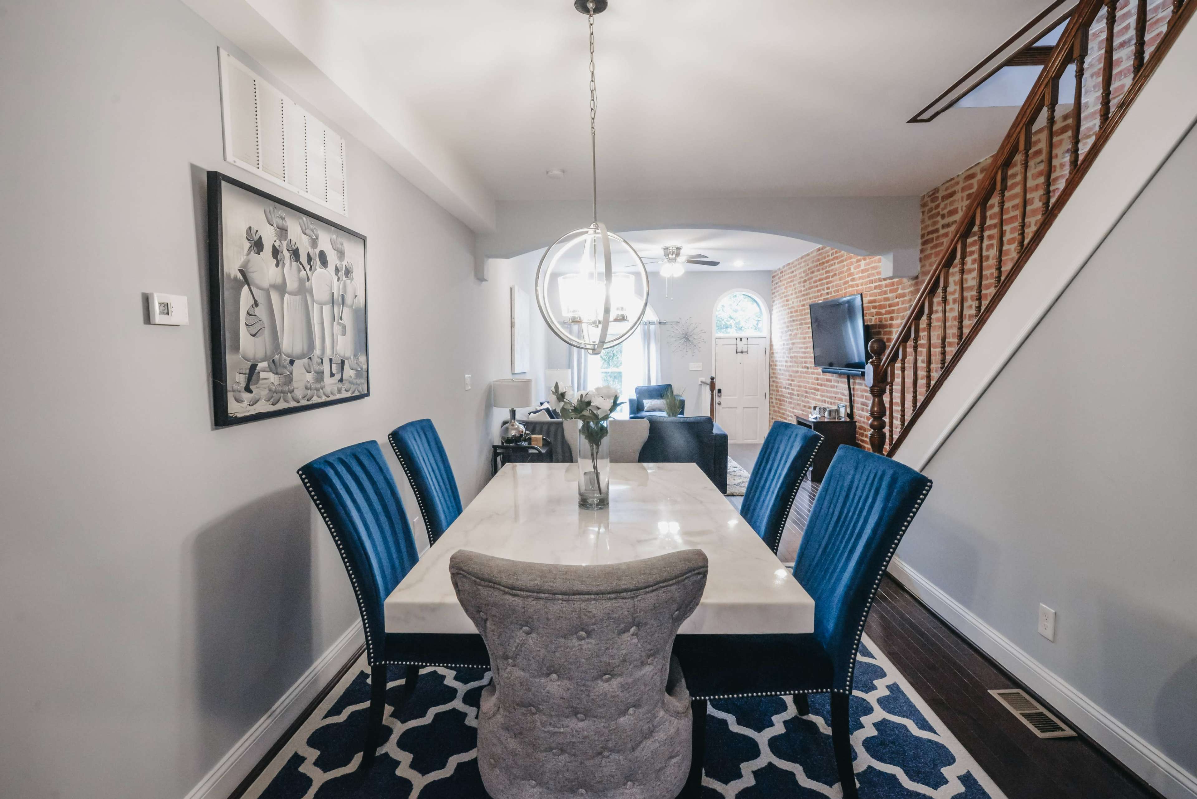 A modern dining area features a marble-top table surrounded by blue upholstered chairs, with a large pendant light overhead and an entrance to a living space in the background.