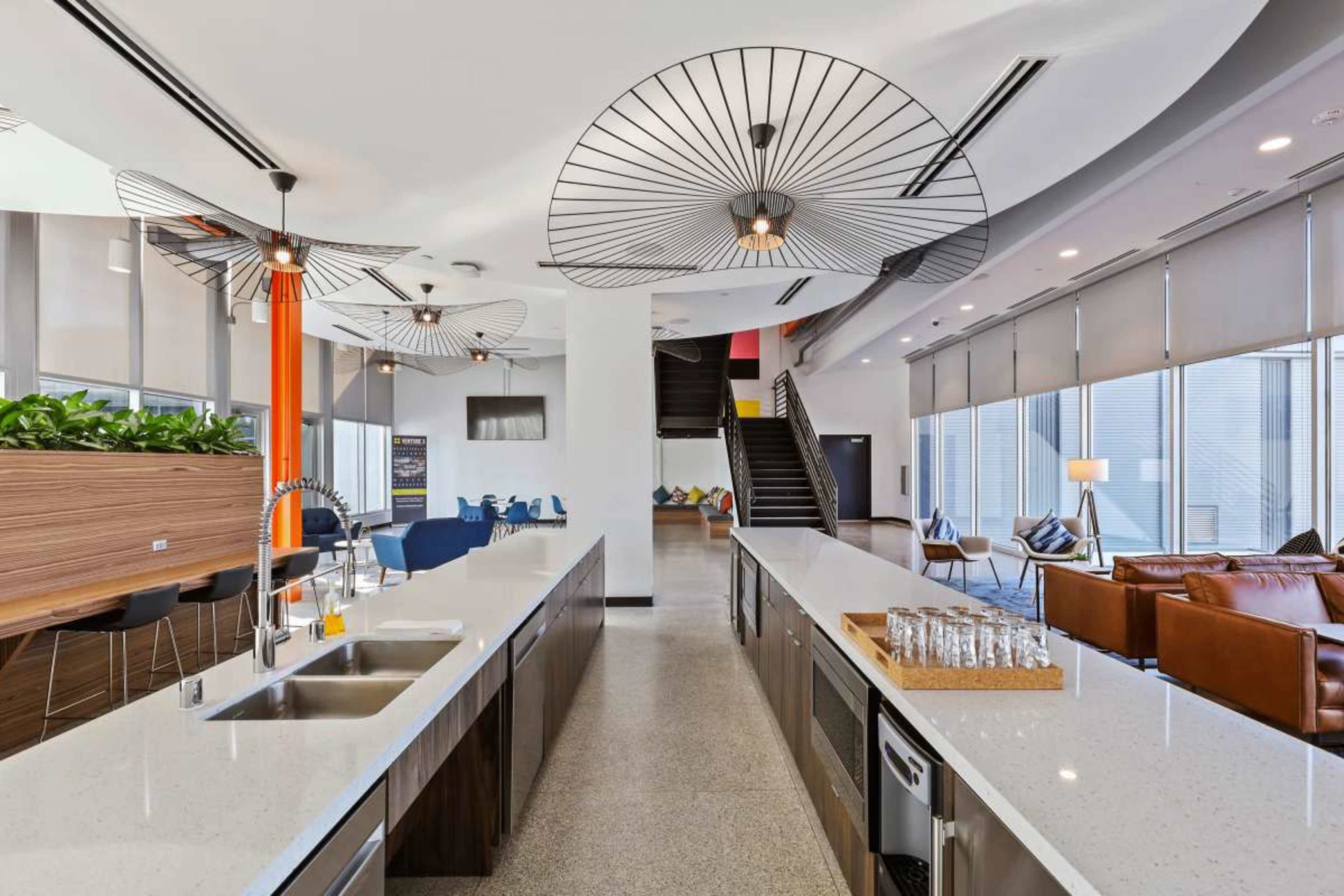 The image shows a modern kitchen area with a long countertop, unique ceiling fixtures, and a seating area with leather couches, all illuminated by natural light from large windows.
