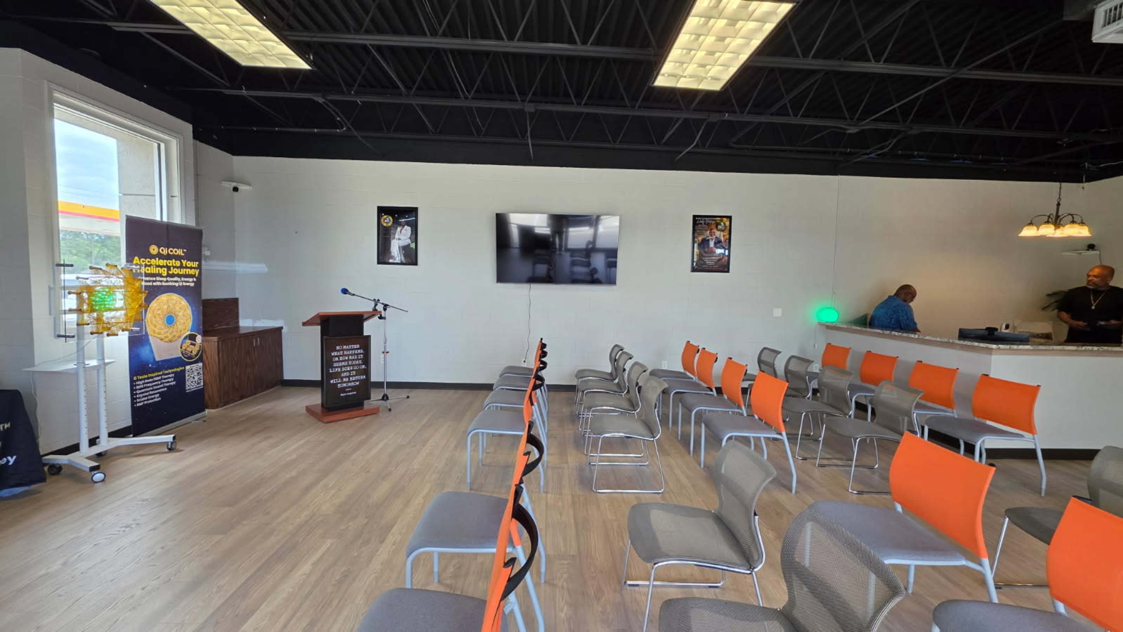 The image shows a seminar room set up with rows of chairs facing a podium, a large screen on the wall, and a bar area in the background.