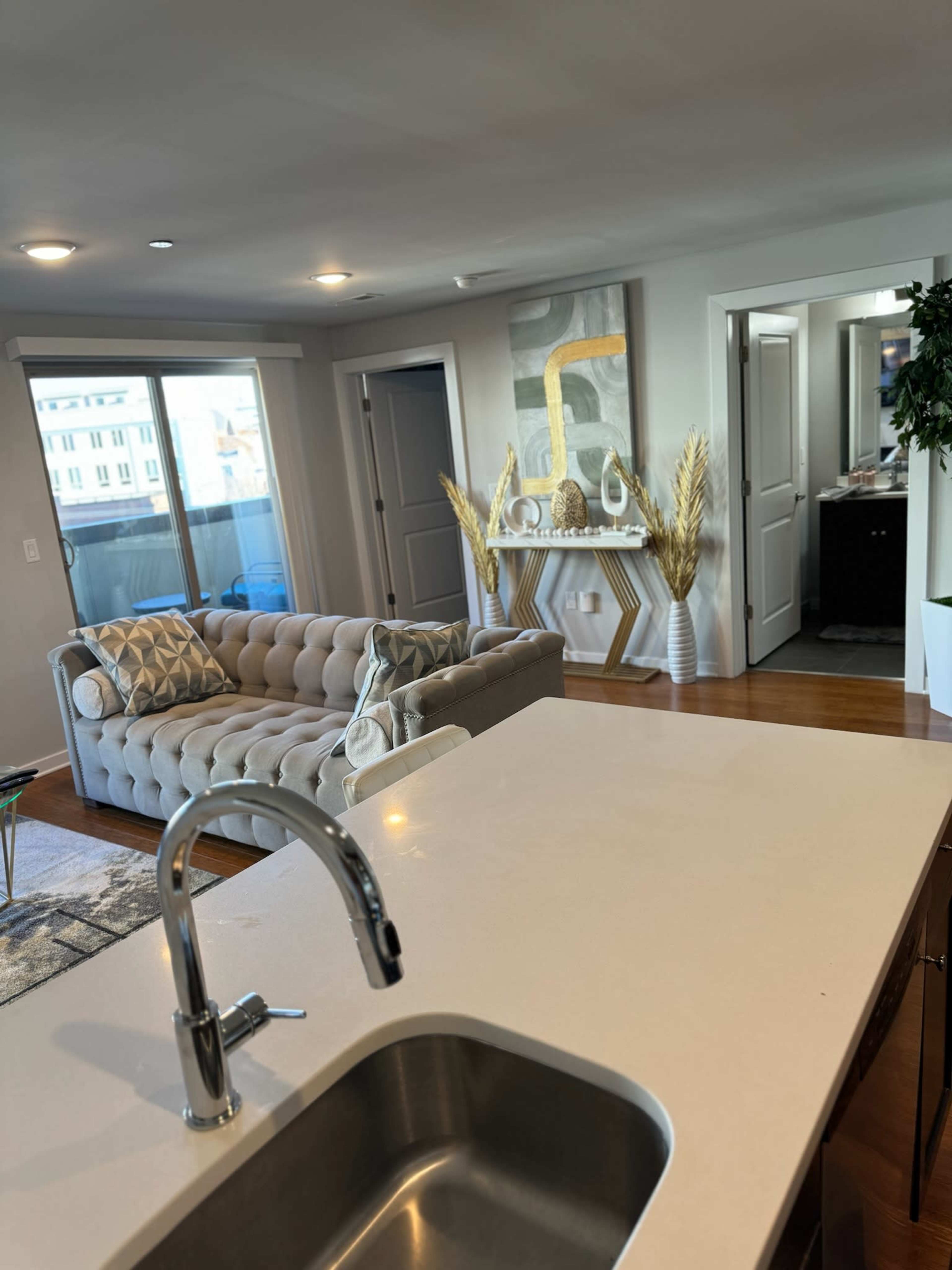 The image shows a modern living space with a kitchen in the foreground, a tufted sofa, and decorative elements in a well-lit room.
