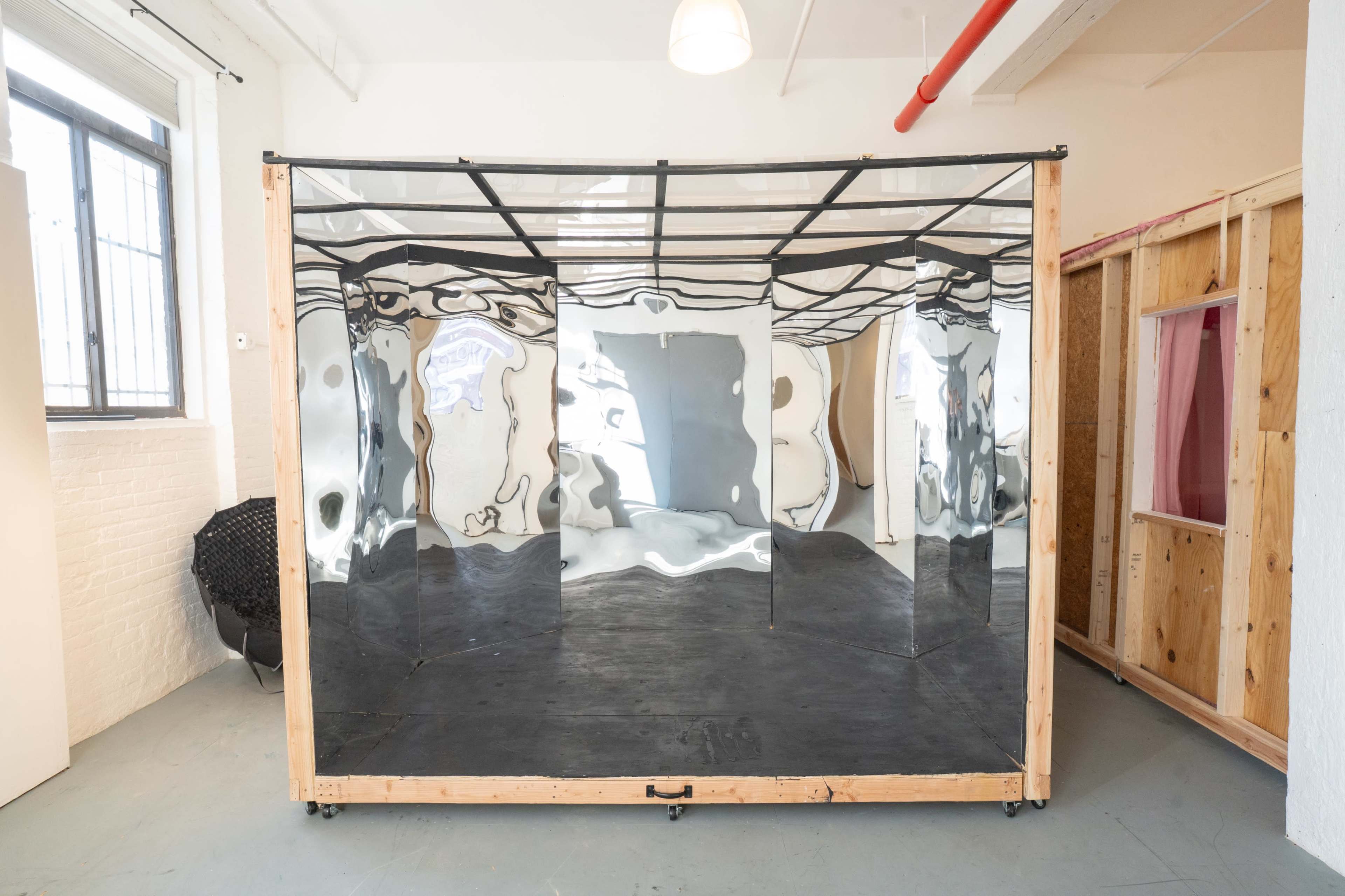 The image shows a large, reflective box structure made of shiny material, situated within a brightly lit, sparse room.