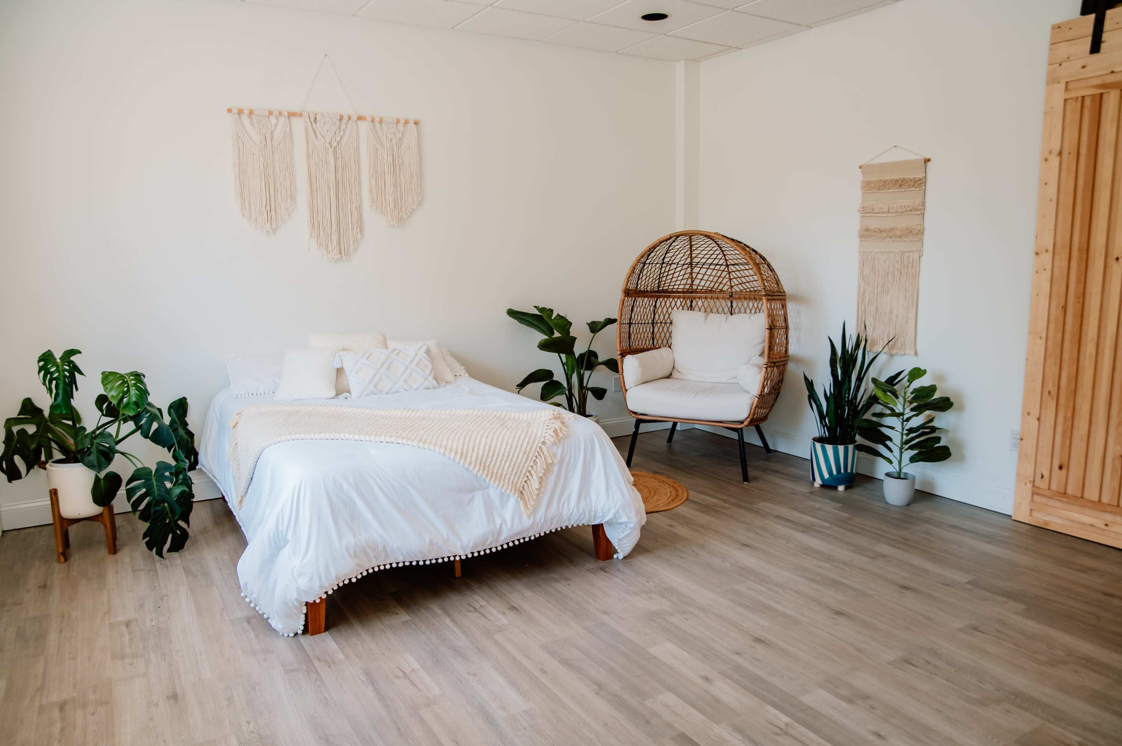 A minimalist bedroom with a bed, potted plants, and a woven chair against a neutral wall.