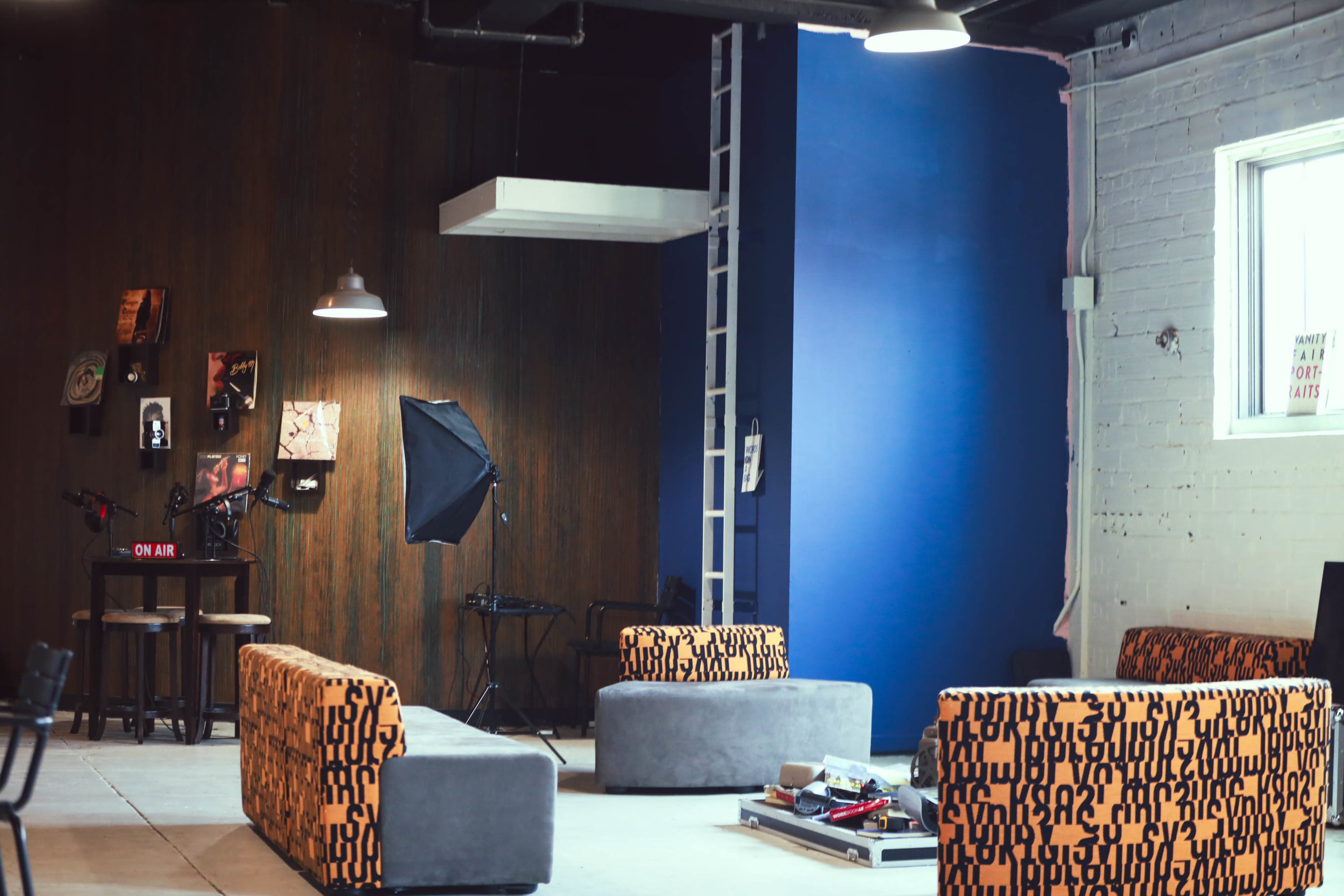 The image shows a modern lounge area with two orange and gray couches, a small table, and a backdrop for photography or filming, accented by industrial lighting.