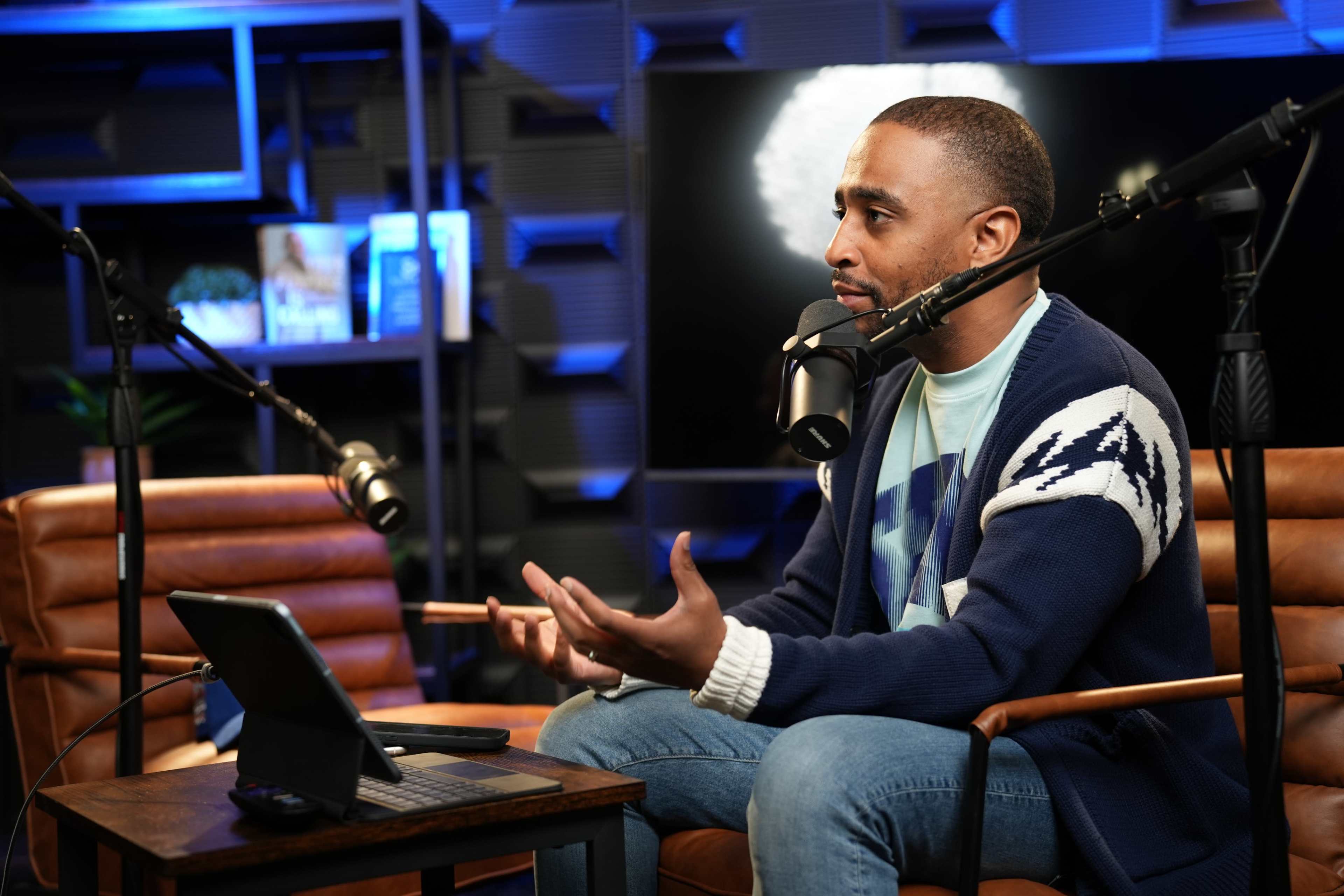 A man is sitting in a modern studio, speaking into a microphone while gesturing with his hands, and a tablet is placed on a table in front of him.