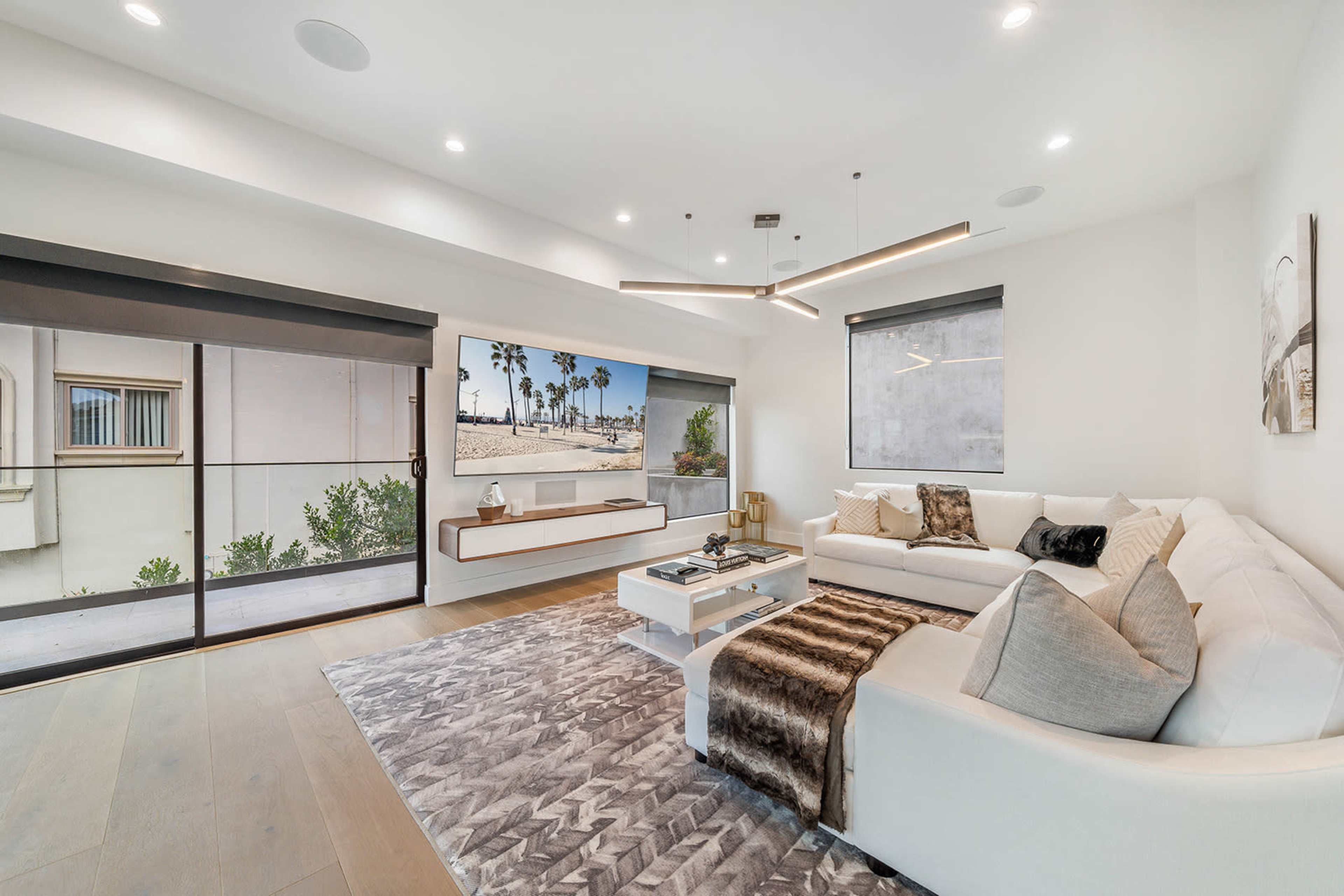 The image shows a modern living room with a large sectional sofa, a coffee table on a patterned rug, and a view of palm trees through a glass sliding door.