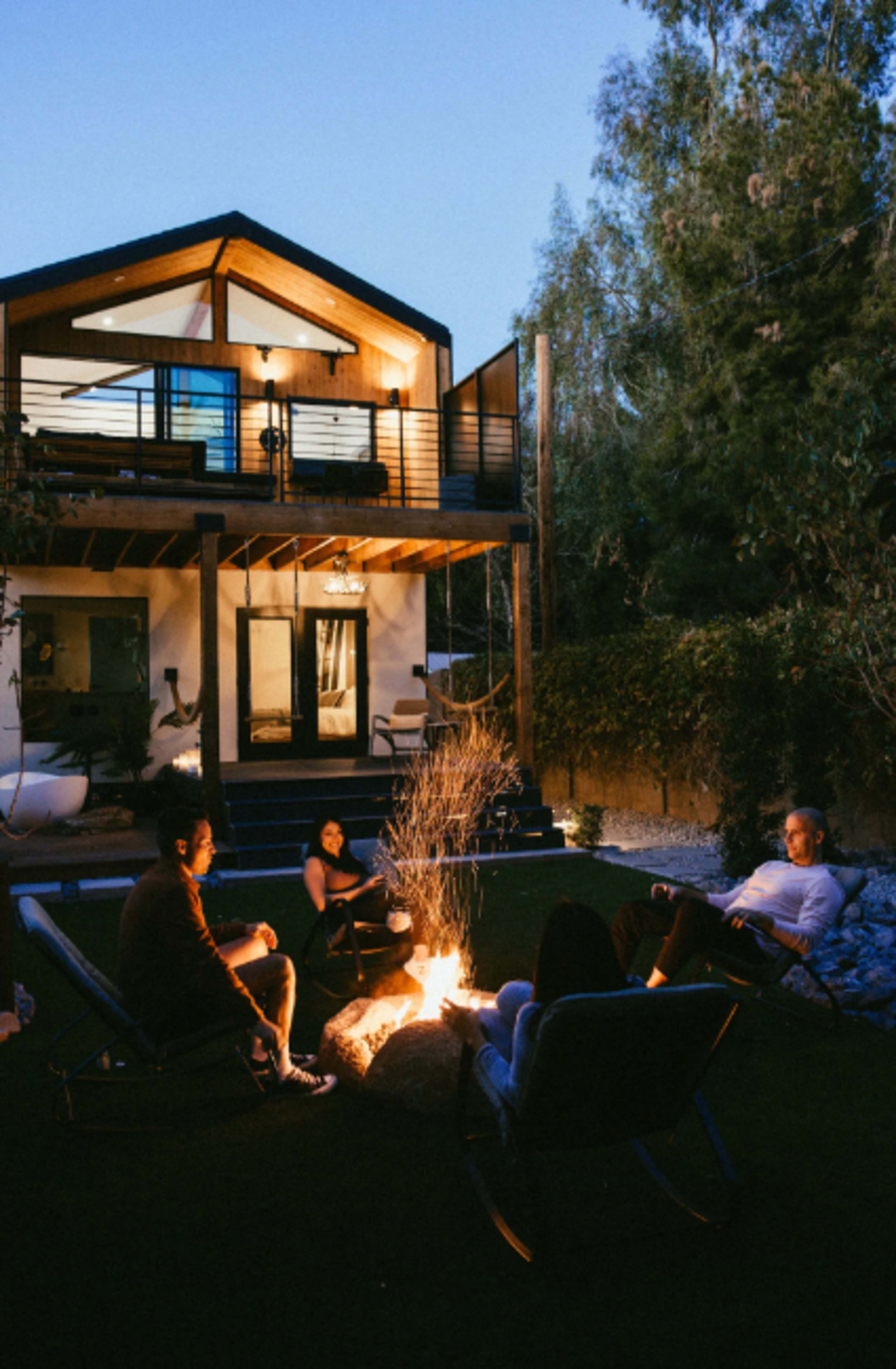 A group of four people sits around a campfire in a backyard, with a modern house visible in the background.