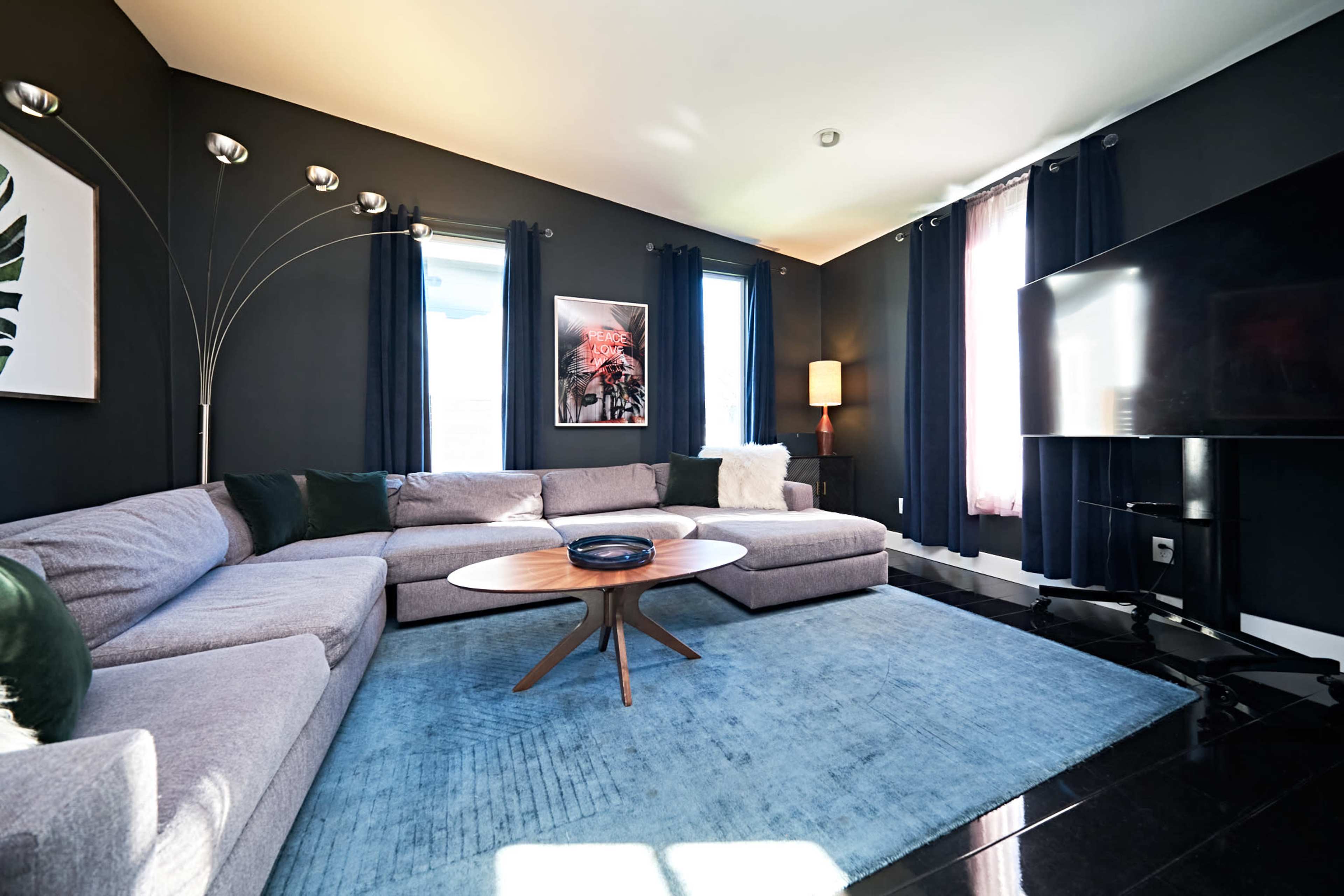 The image shows a modern living room featuring a large sectional couch, a round coffee table, a flat-screen TV, and large windows with black curtains.