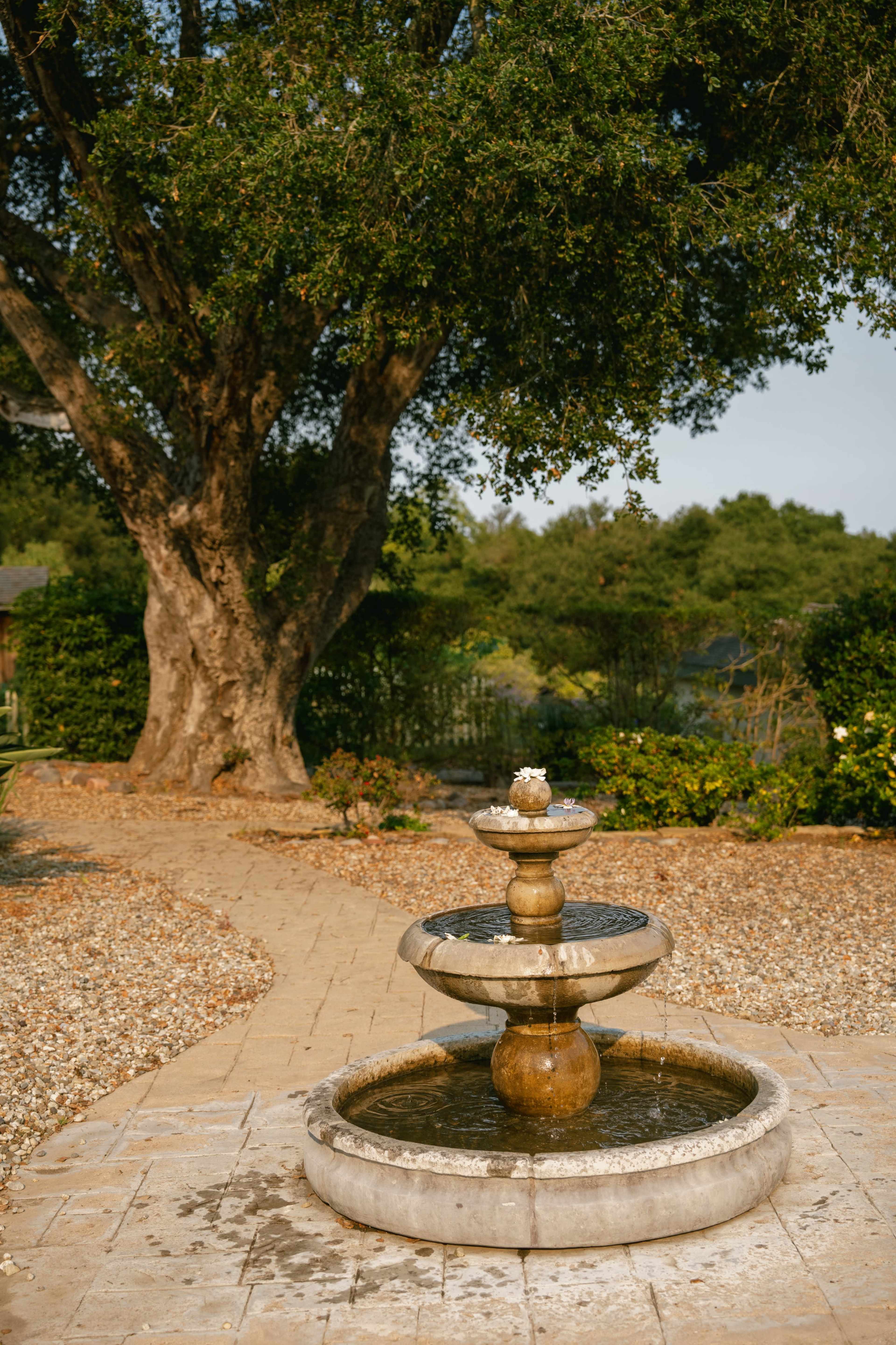 A stone fountain with three tiers is set in a landscaped garden, surrounded by greenery and a winding path.