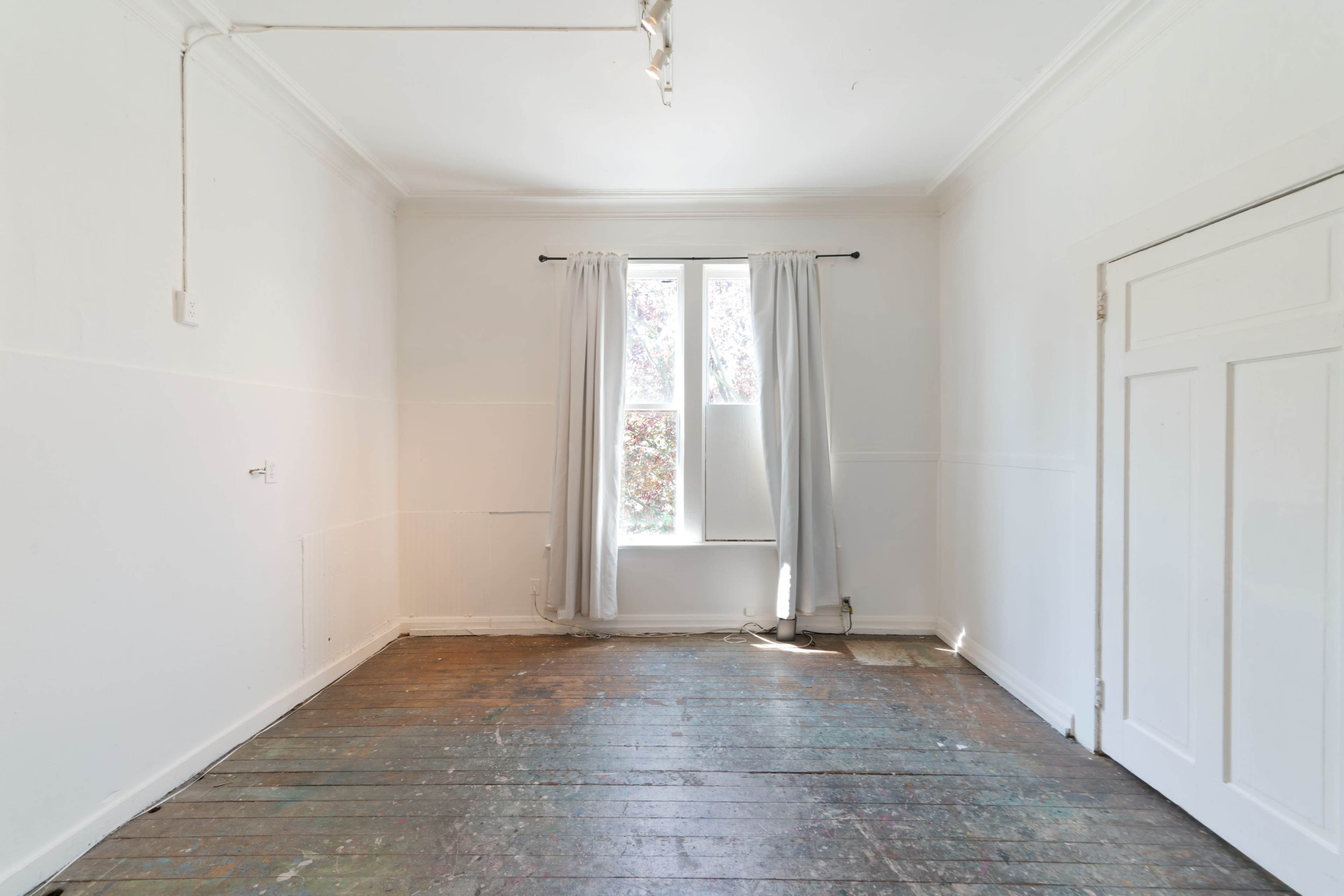 A sparsely furnished room features a single window with white curtains, wooden floors, and a door on one side.