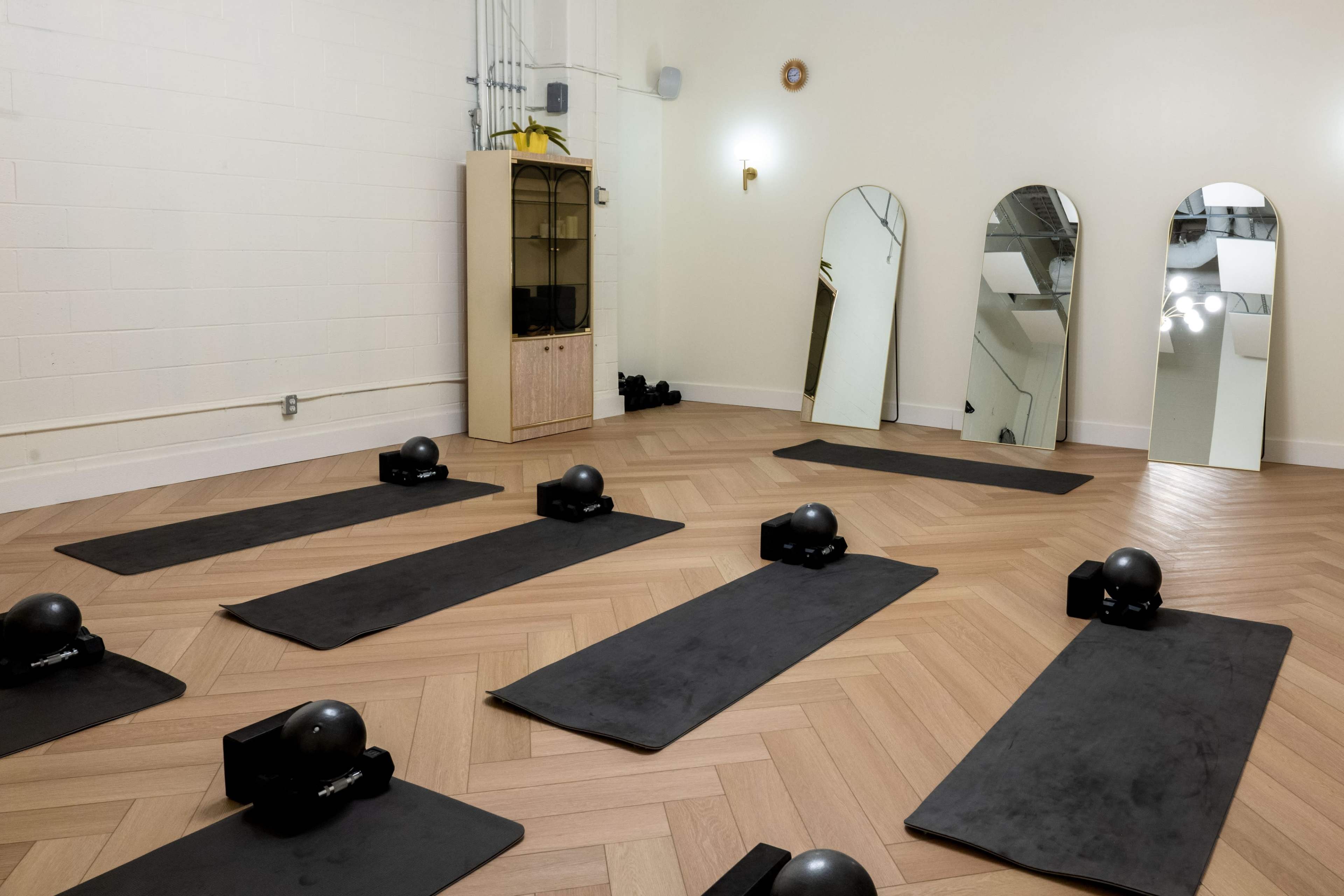 The image shows a yoga or fitness studio featuring several workout mats arranged on a hardwood floor, with large mirrors and a cabinet in the background.