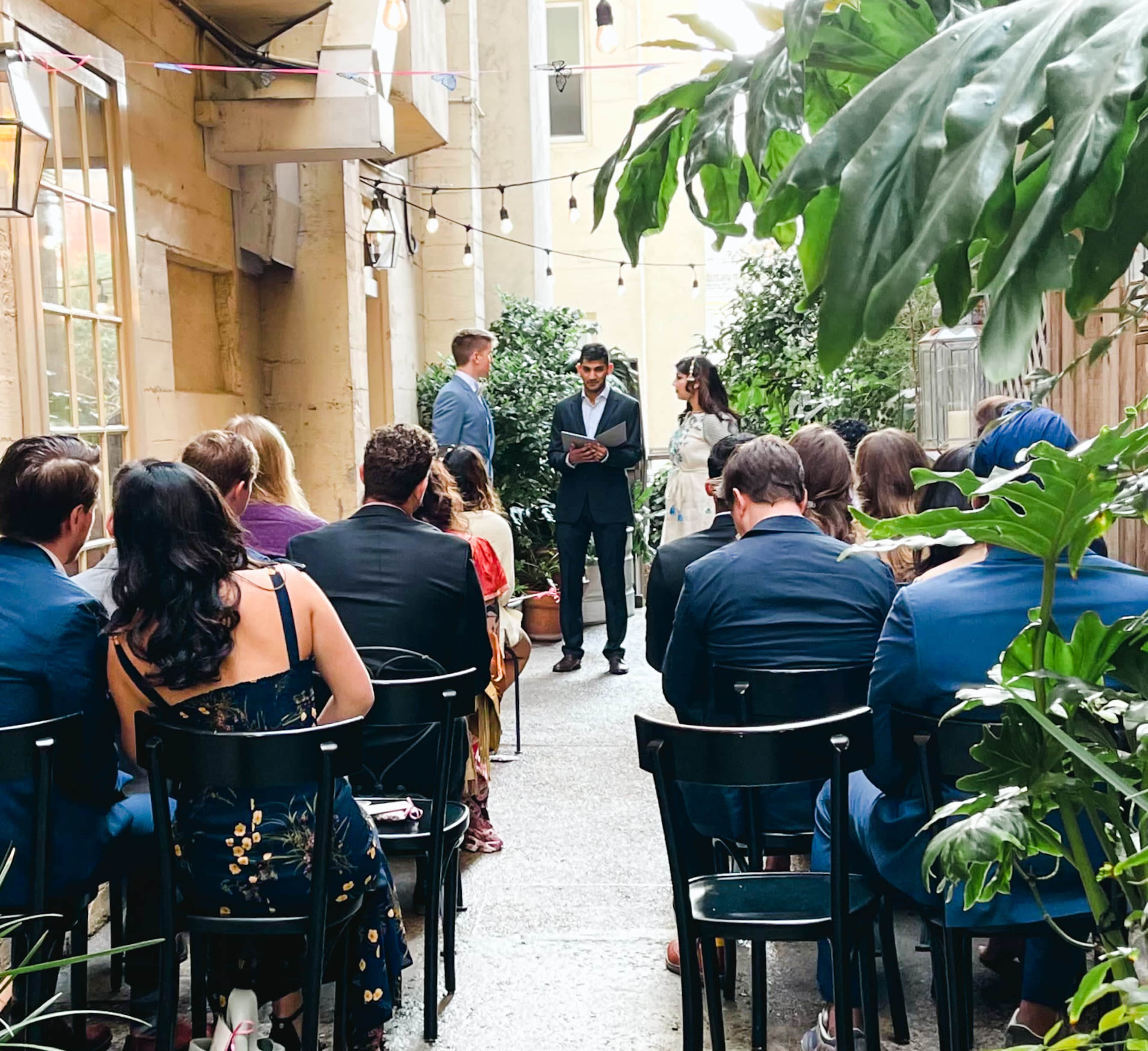 A couple stands at the front of an outdoor venue, surrounded by guests seated in chairs, while an officiant appears to be conducting a ceremony.