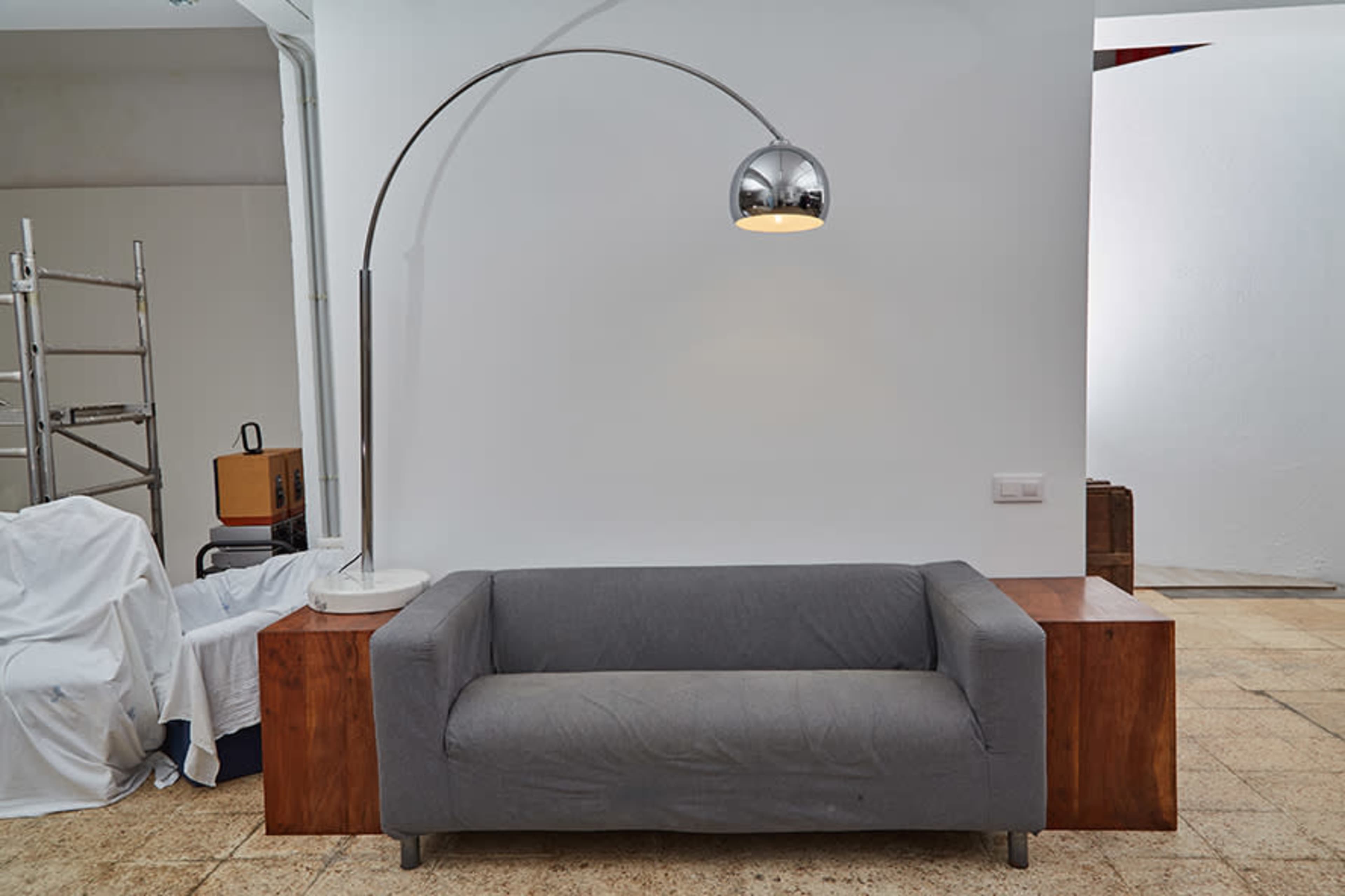 A grey sofa is positioned against a white wall, with a tall arc floor lamp beside it and wooden furniture elements in the background.