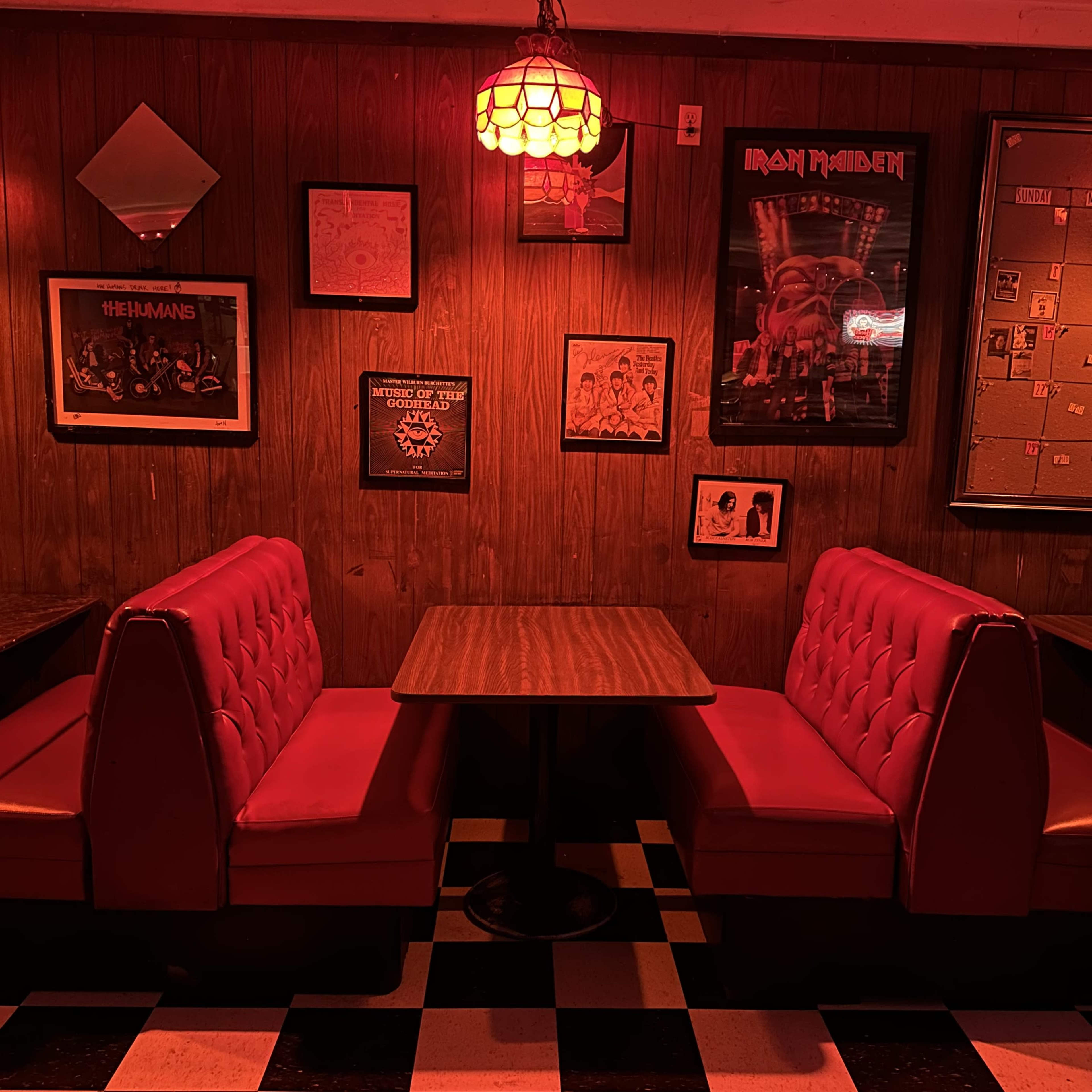 A cozy diner booth with red cushioned seating and a wooden table under dim red lighting, surrounded by framed posters and artwork on wooden-panelled walls.