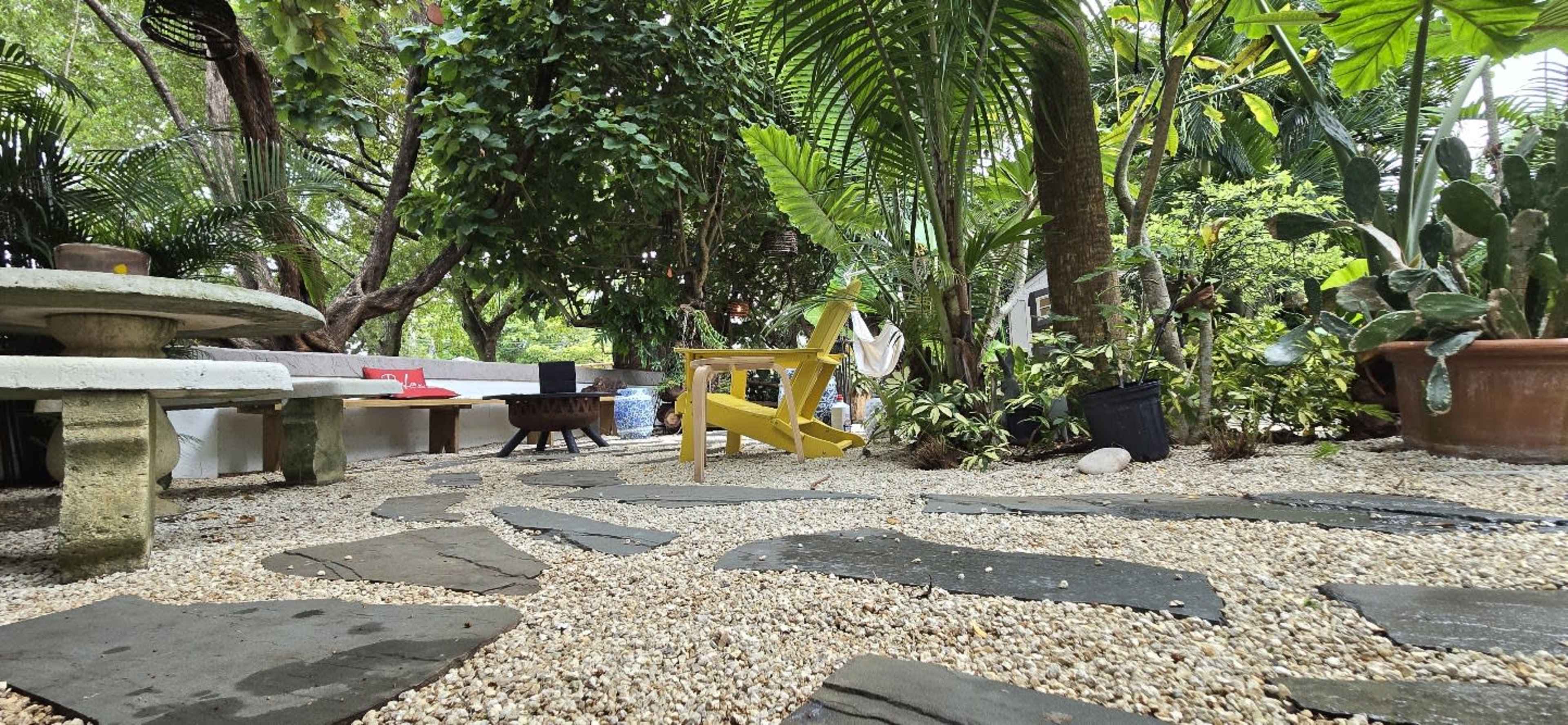 The image shows a garden area featuring a stone pathway, a yellow chair, and various plants surrounded by trees.