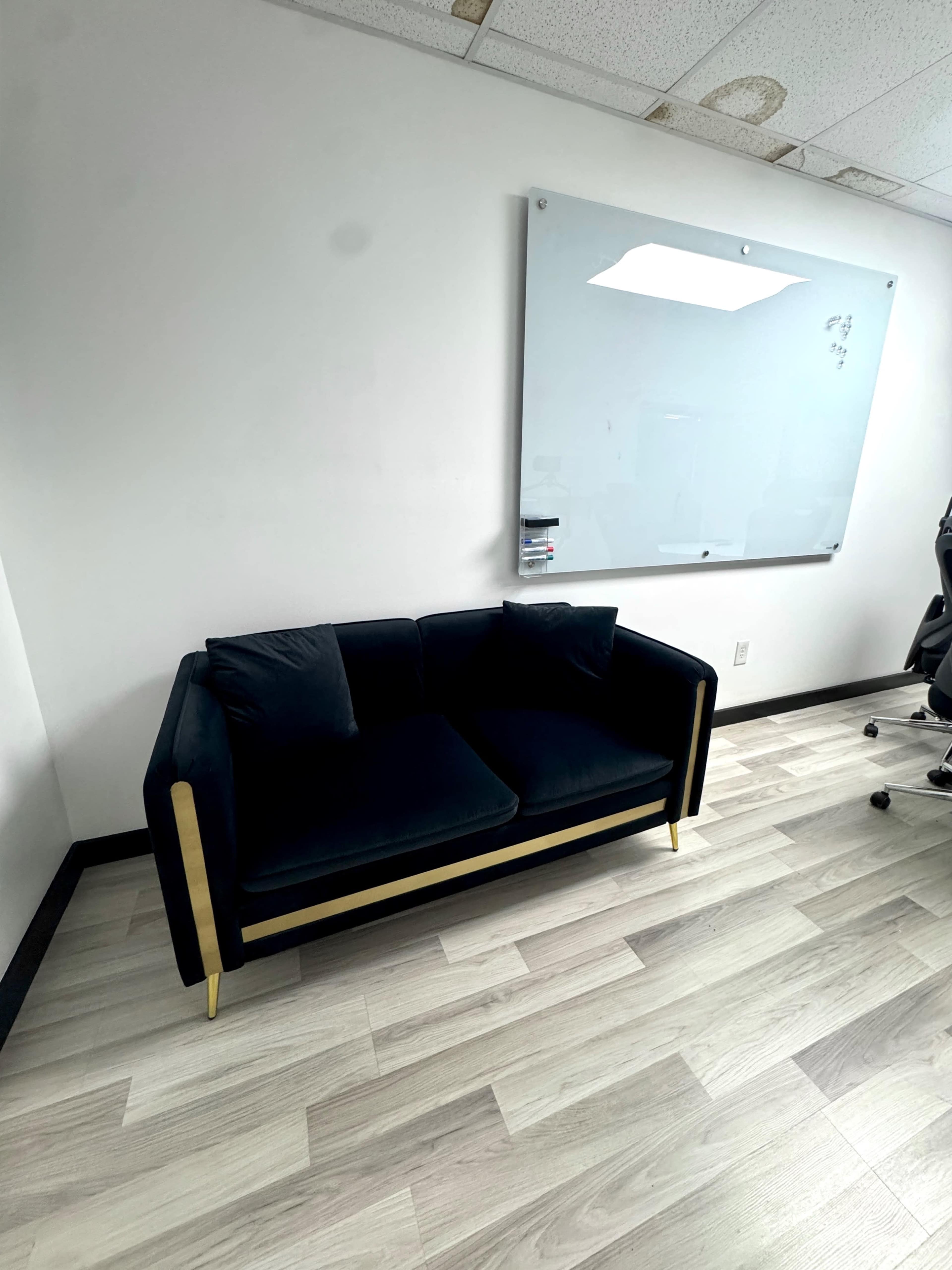 The image shows a dark-colored sofa with gold accents positioned near a whiteboard in a modern office space.