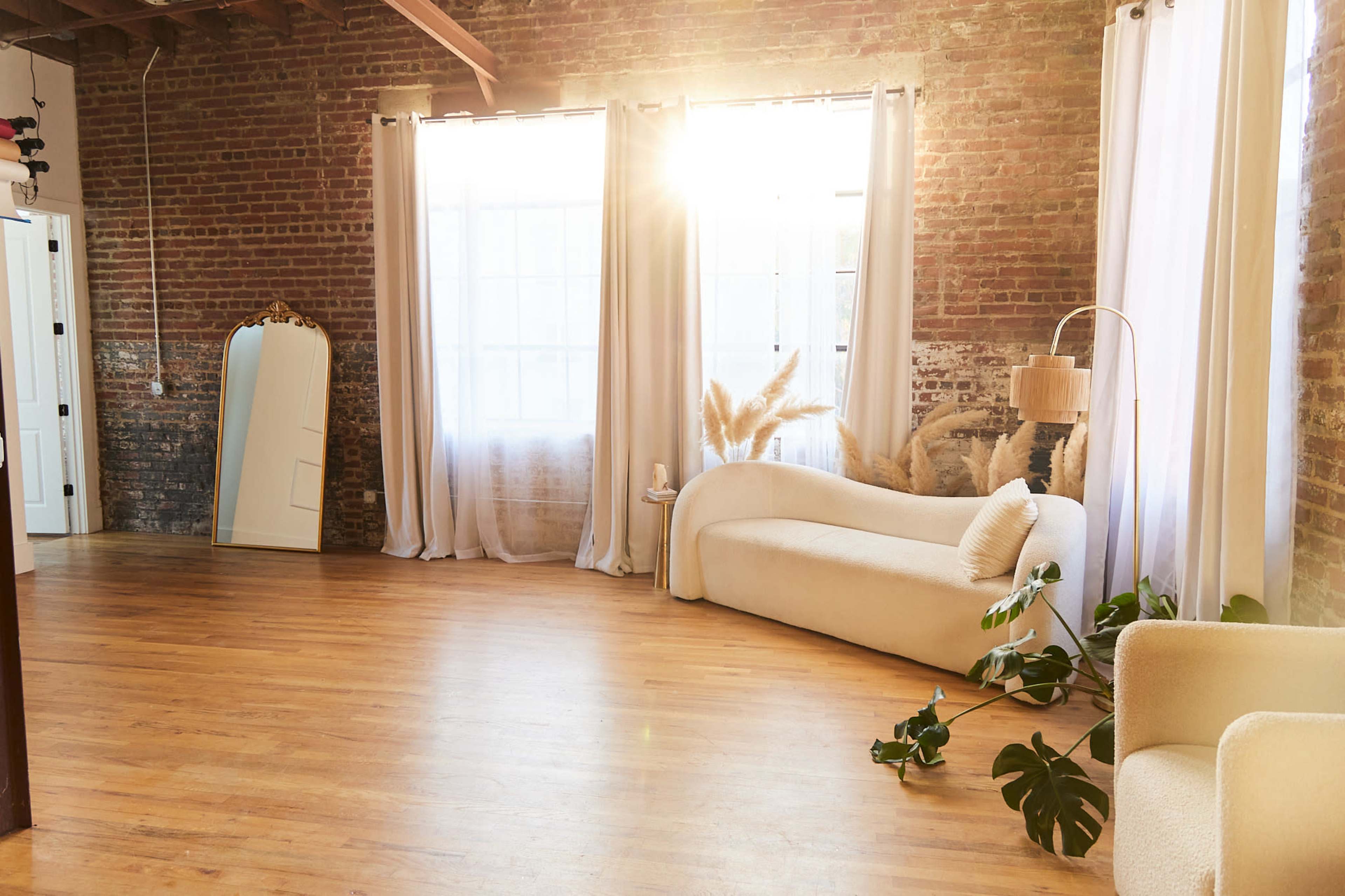 A sunlit interior features a spacious room with exposed brick walls, large windows, and minimalist furniture, including a white sofa and potted plants.