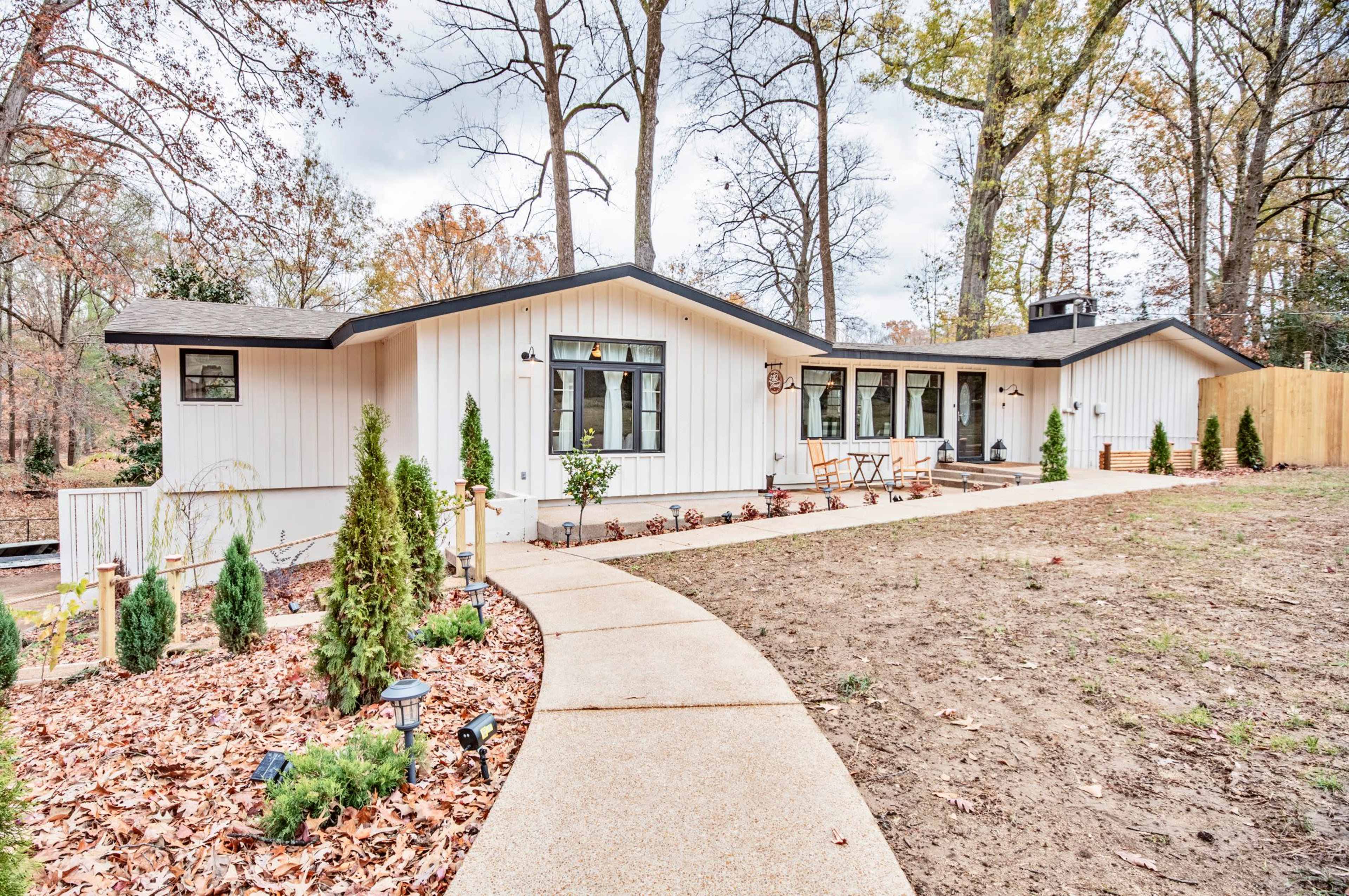 A modern single-story house with a curved walkway, surrounded by landscaped shrubs and trees.