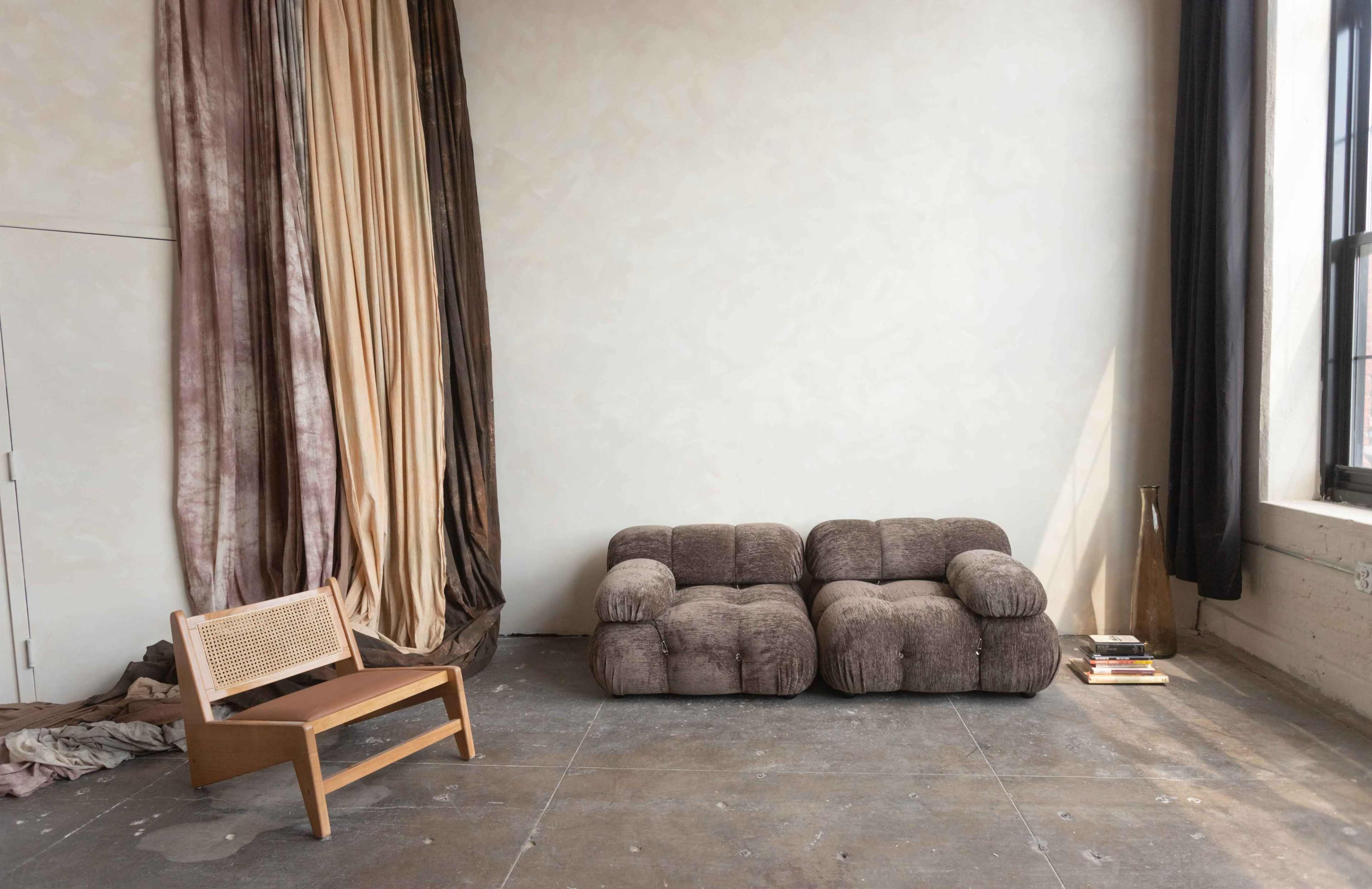 A minimalistic interior space with a brown fabric couch, a wooden chair, and draped fabric in various colors against a wall.
