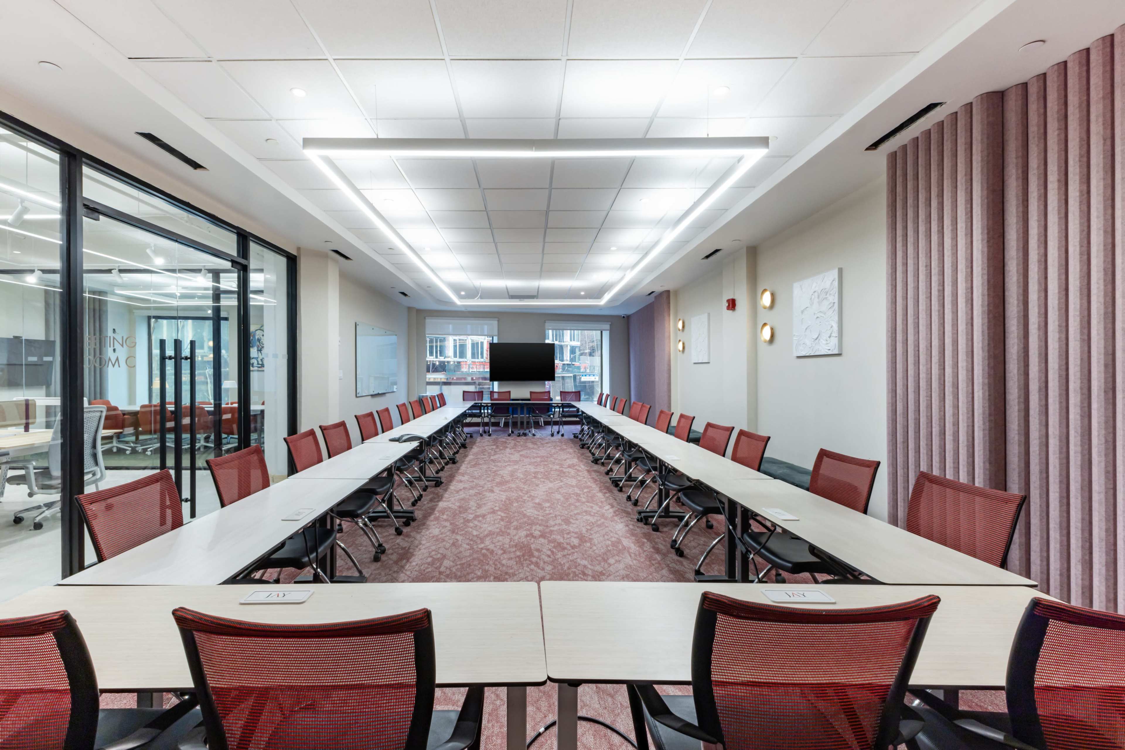 A modern conference room features a long rectangular table arranged with red chairs, flanked by glass-walled meeting spaces and adorned with muted pink fabric wall panels.