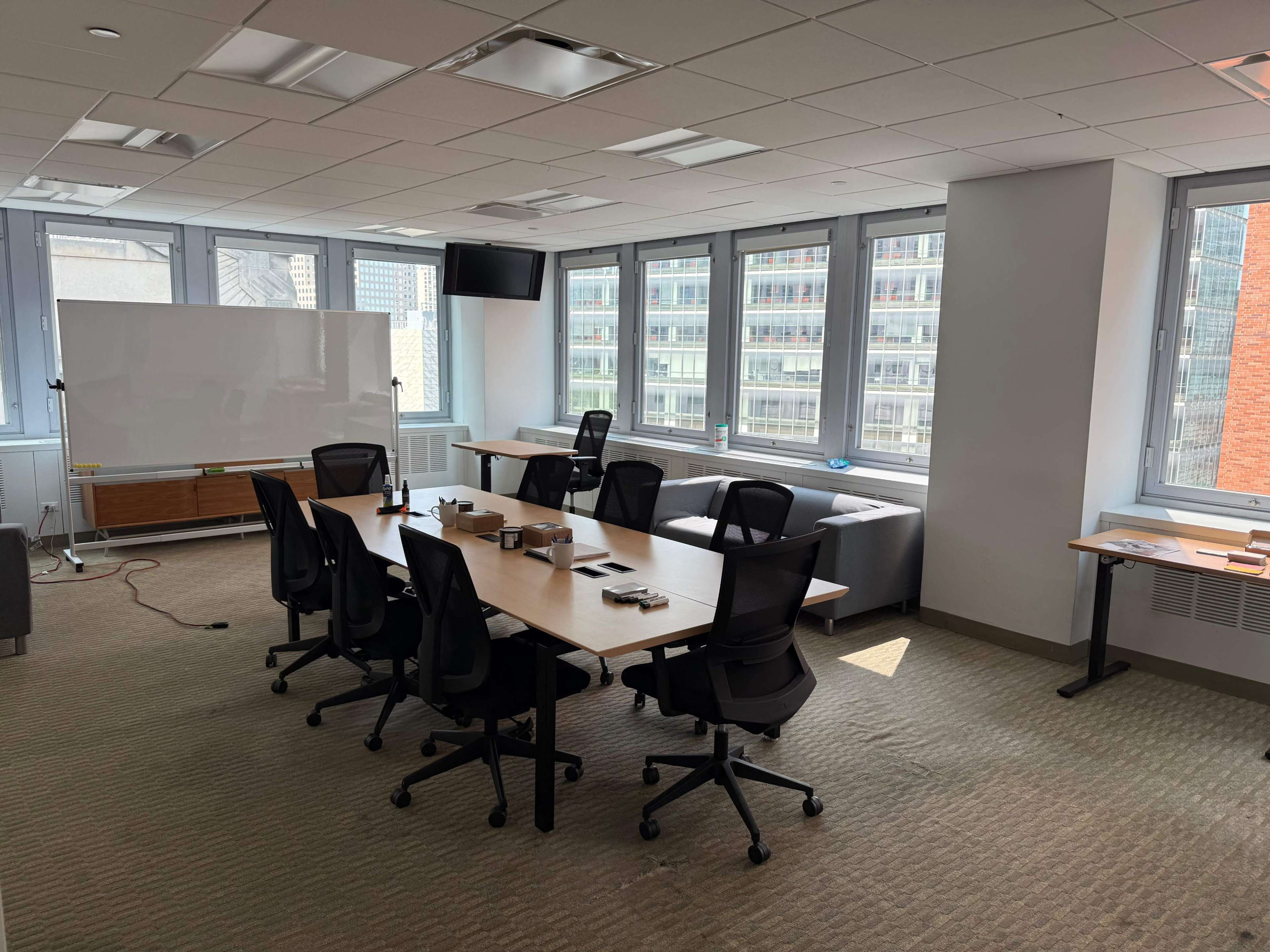 A conference room is set up with a long table surrounded by chairs, a whiteboard, and large windows overlooking a cityscape.