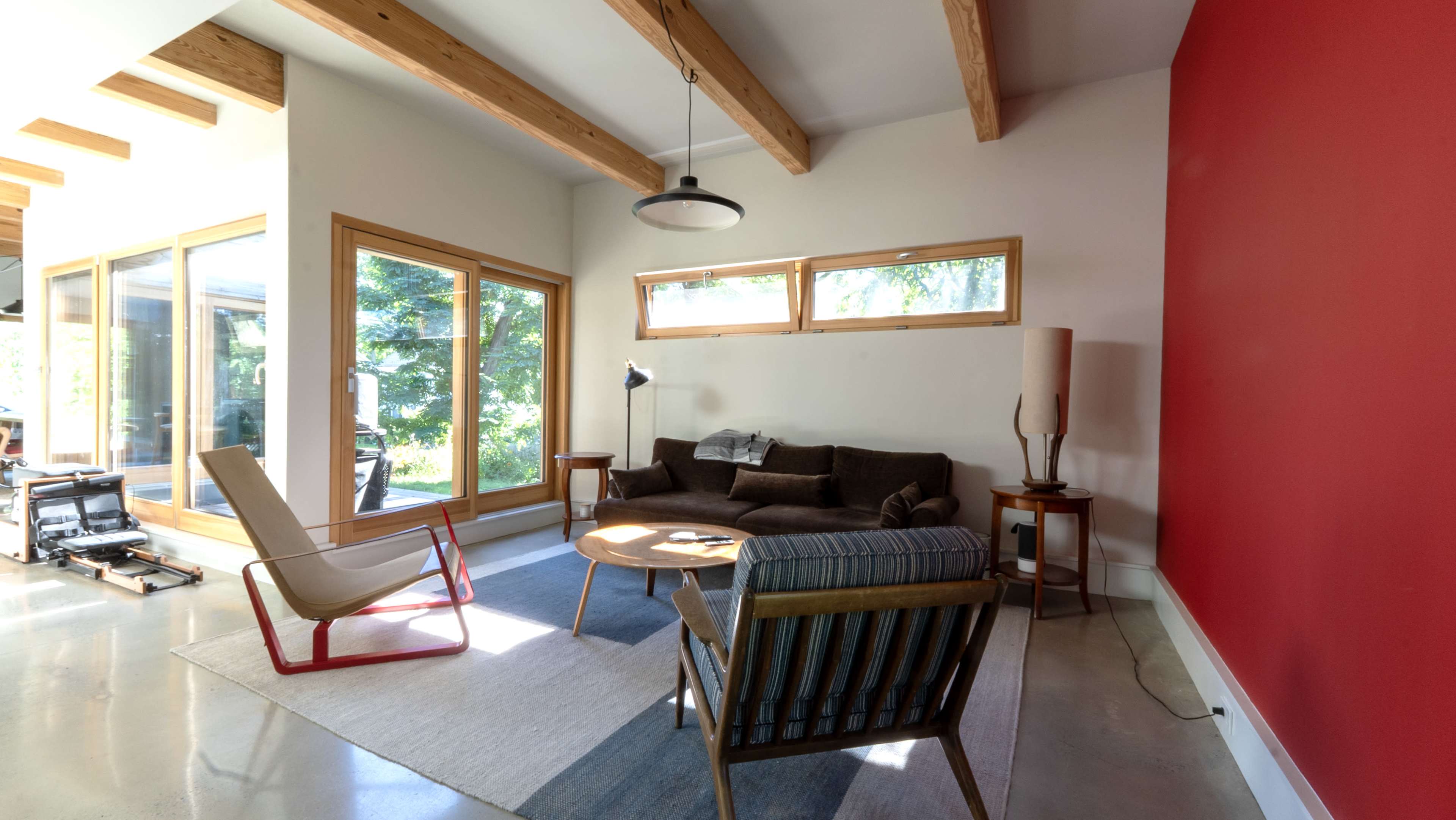 The image shows a modern living room with wooden beams, large windows, and minimalistic furniture, including a couch, a chair, and a round table.
