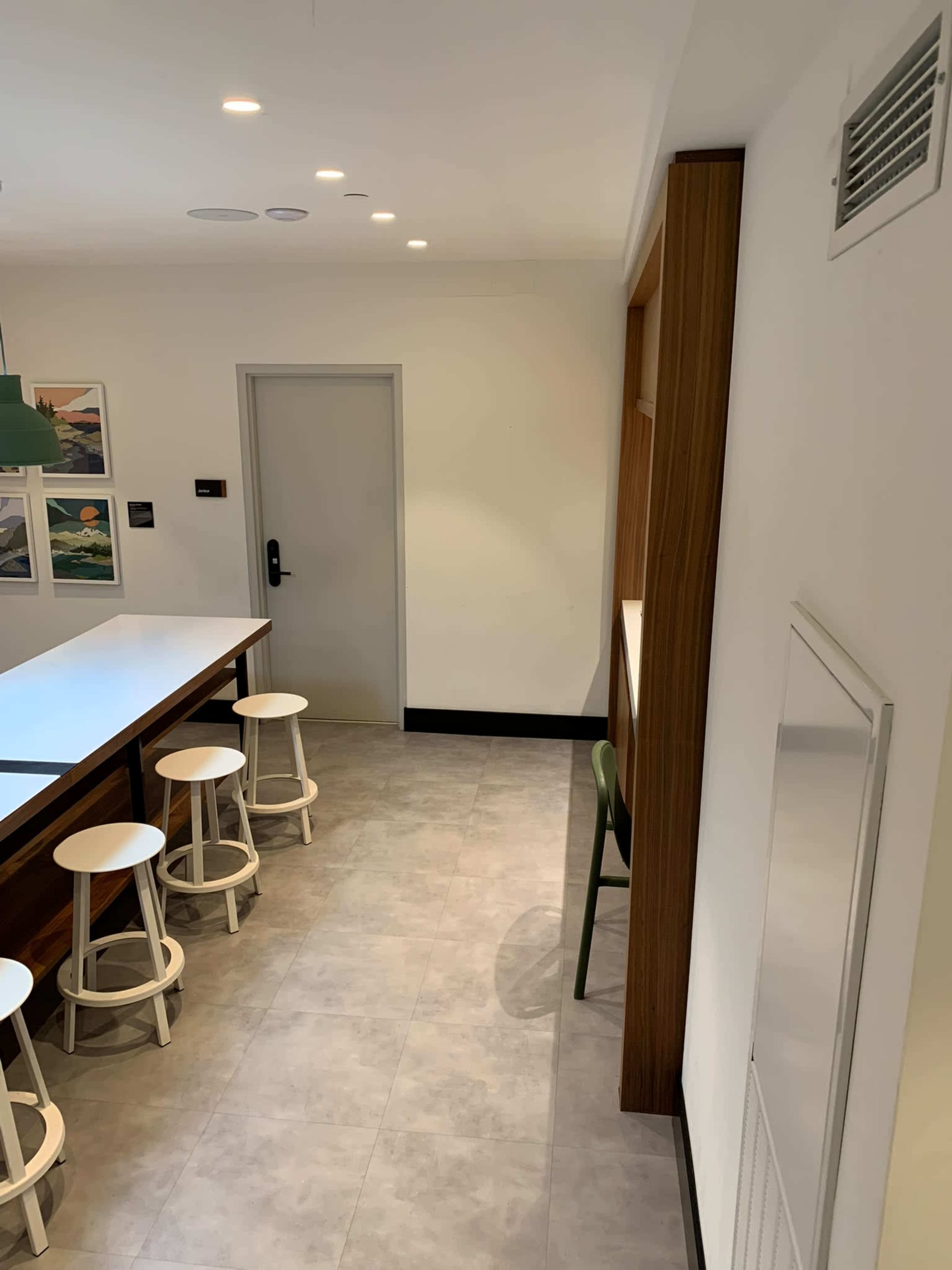 The image shows a modern kitchen area with a long white countertop and several white stools, featuring a doorway and wall-mounted shelves.