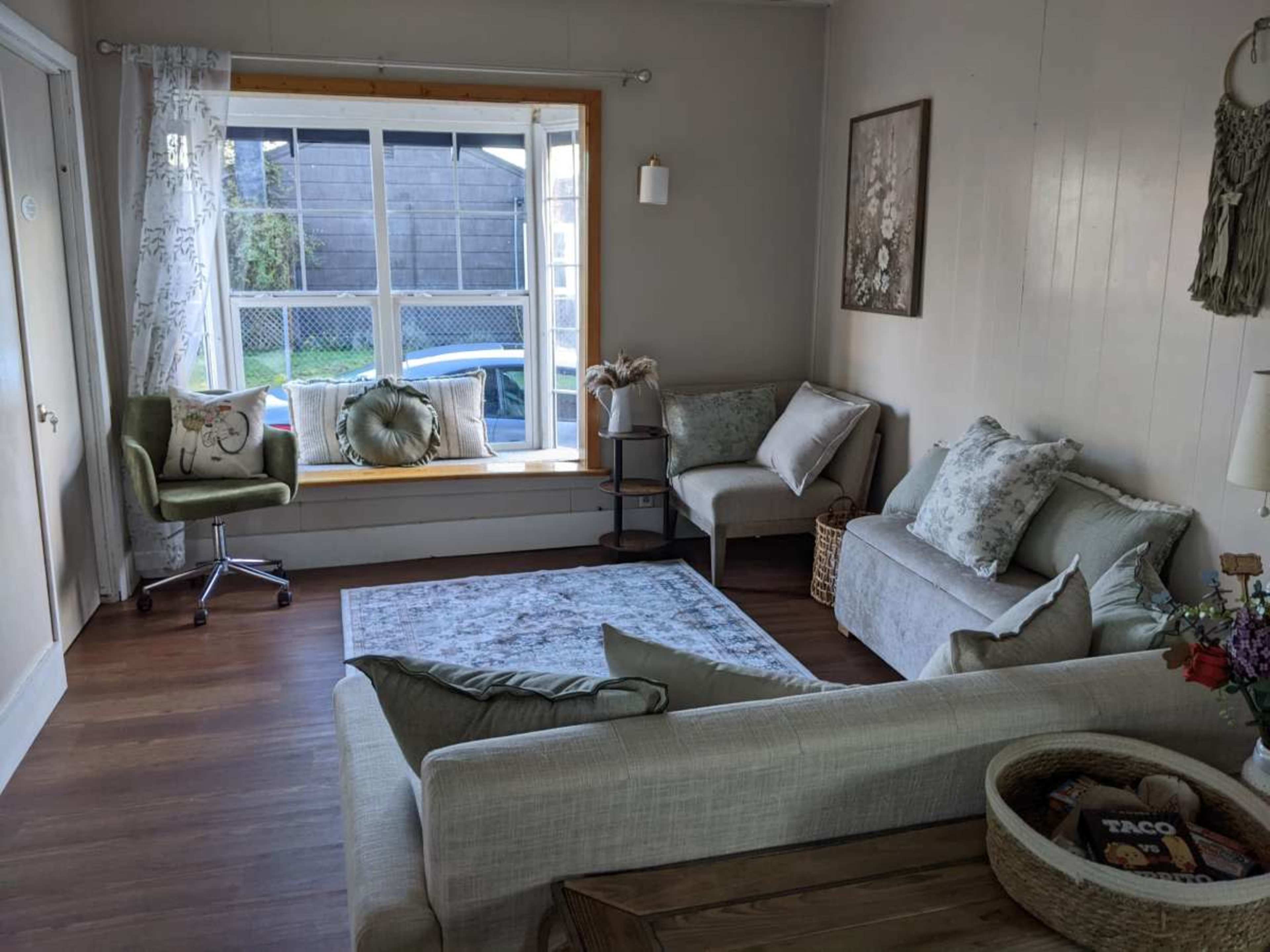 The image shows a cozy living room with a large window, a light-colored sofa, a decorative armchair, and a rug on a wooden floor.