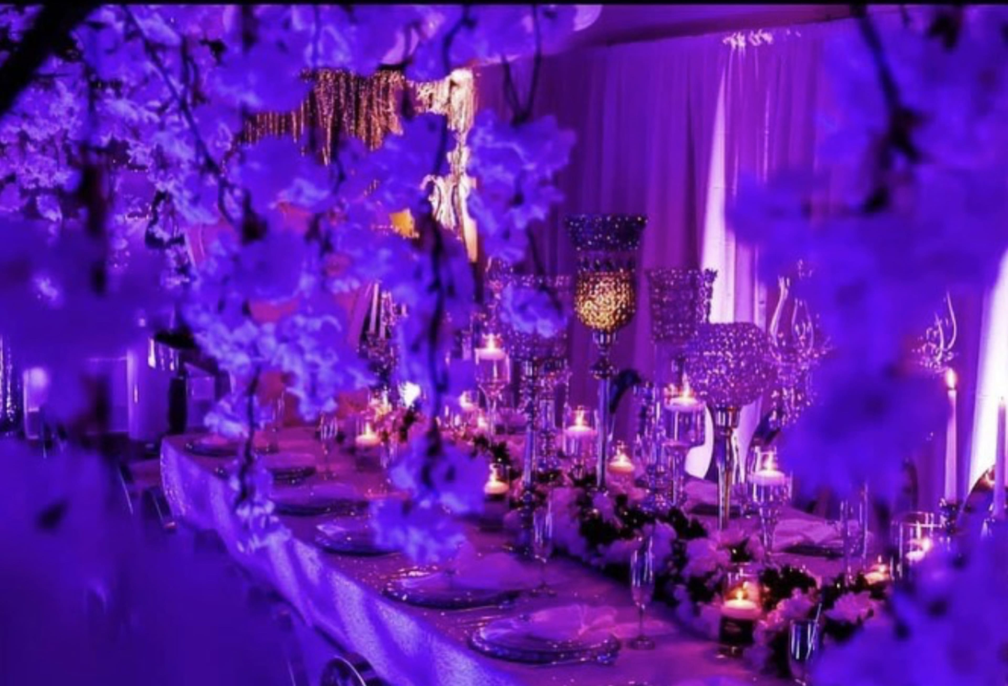 A banquet table is elegantly set with candles and floral arrangements, surrounded by purple-lit decor.