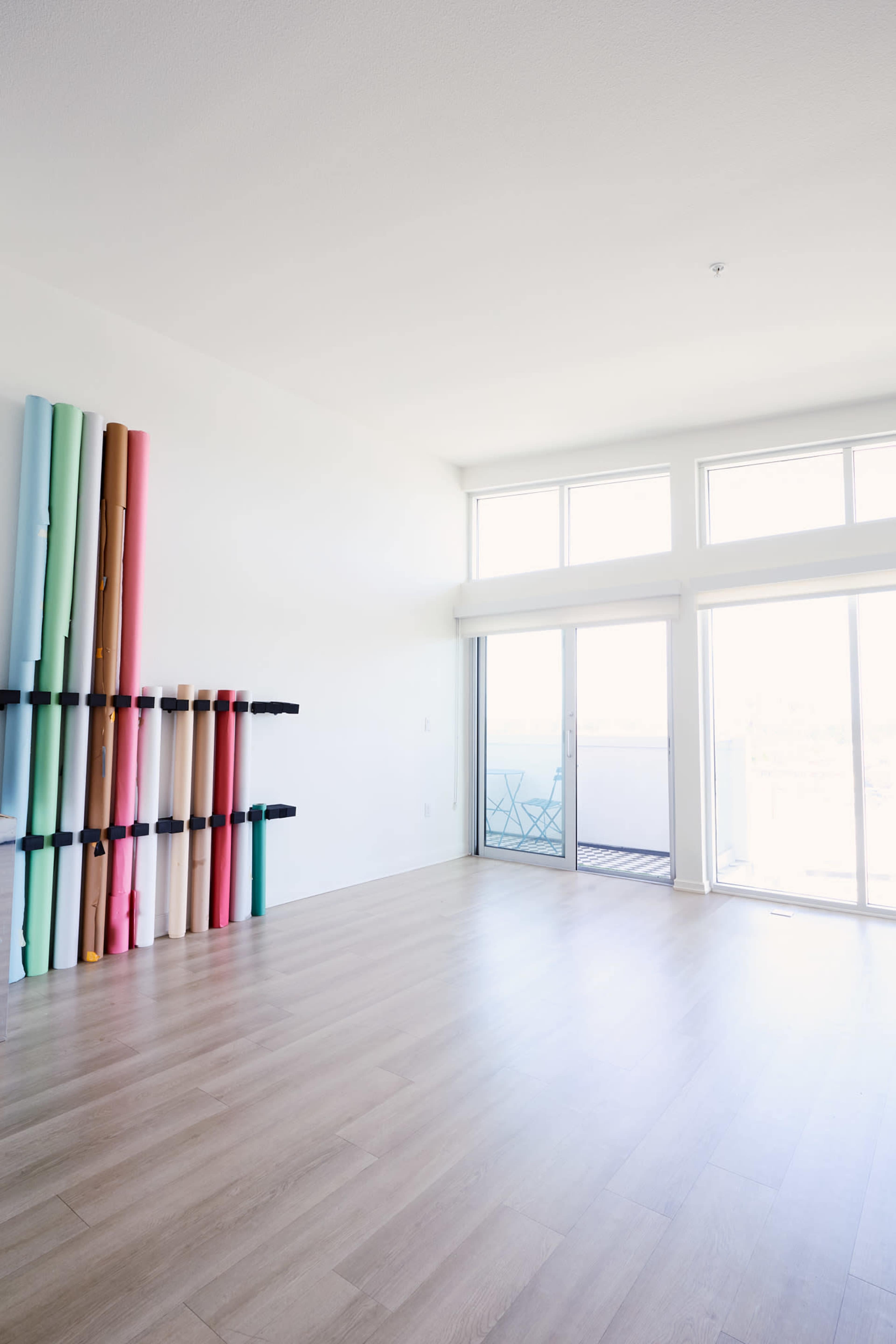 The image shows a bright, empty room with large windows and a wall-mounted rack holding rolled materials in various colors.