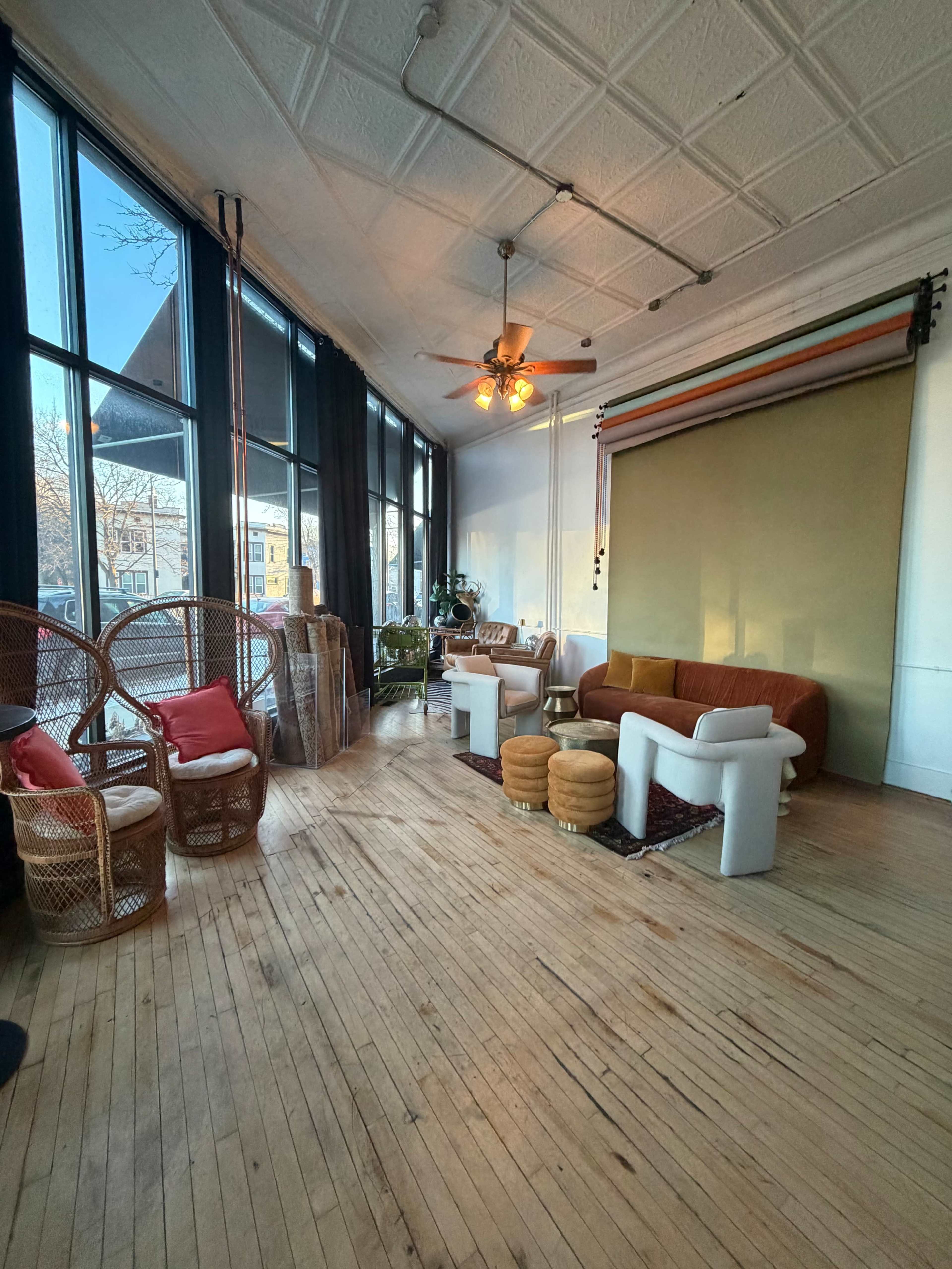 The image shows a spacious room with wooden floors, large windows, and a mix of seating including wicker chairs and a sofa.